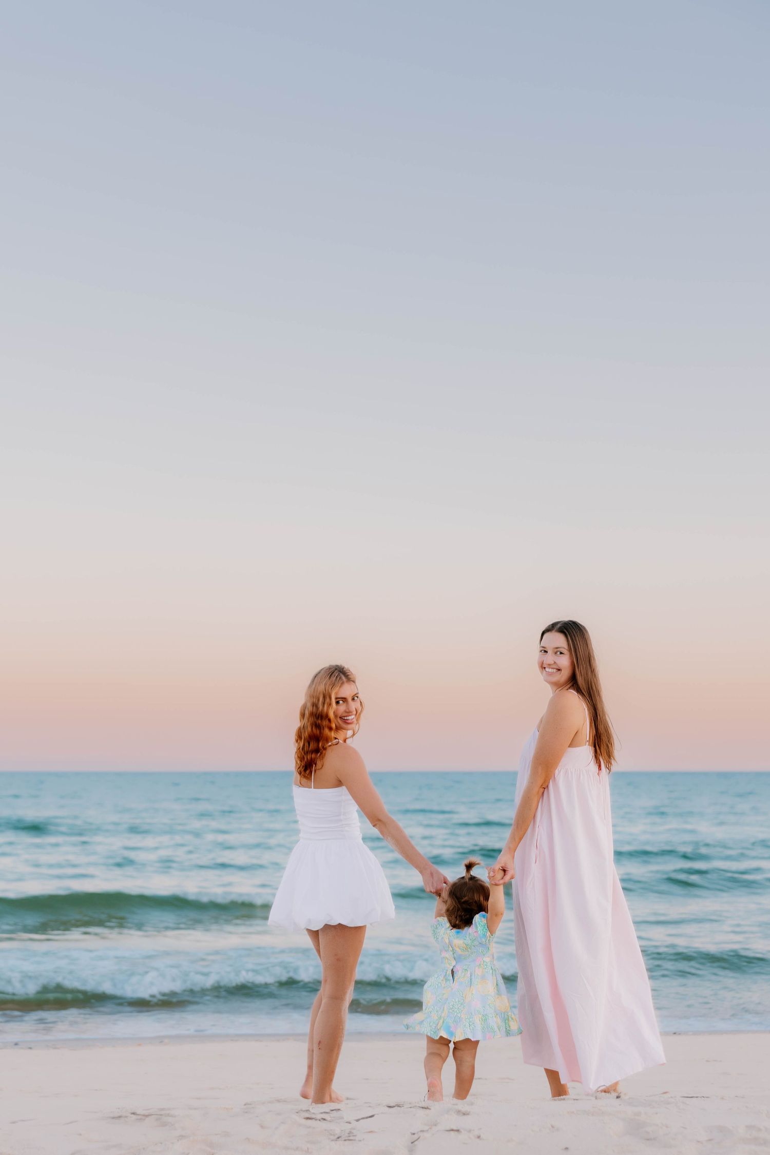 Two women in white and pink dresses hold hands with a toddler on a sandy beach at sunset.