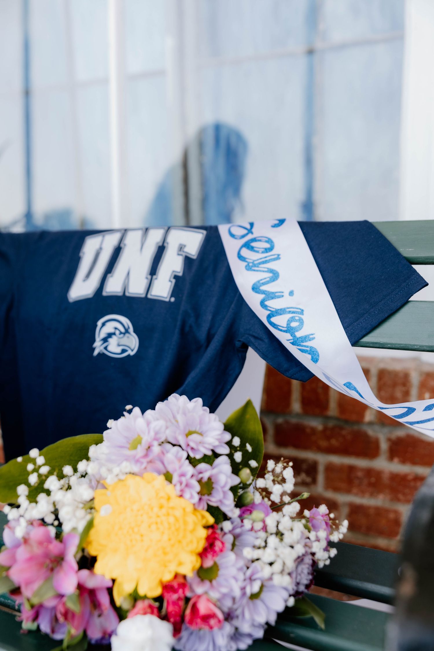 UNF navy blue college sweatshirt beside a colorful bouquet of flowers with a white sash on a bench.