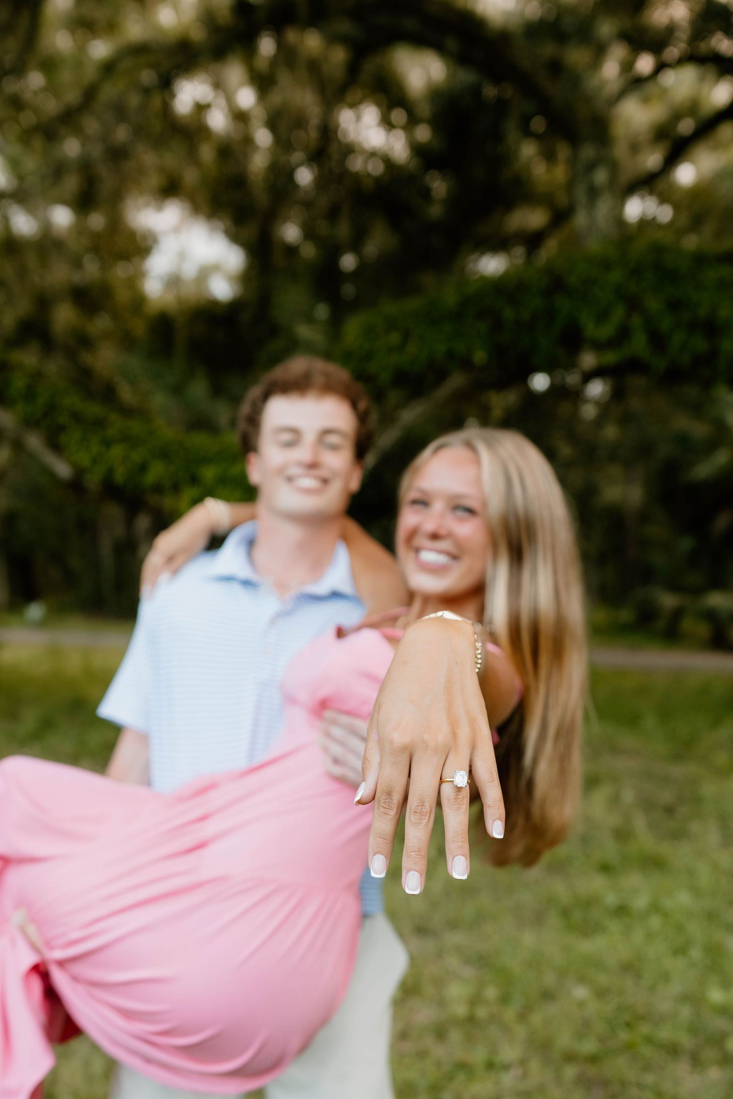 A joyful couple celebrates their engagement outdoors, woman showing diamond ring while being carried by smiling man in blue shirt.