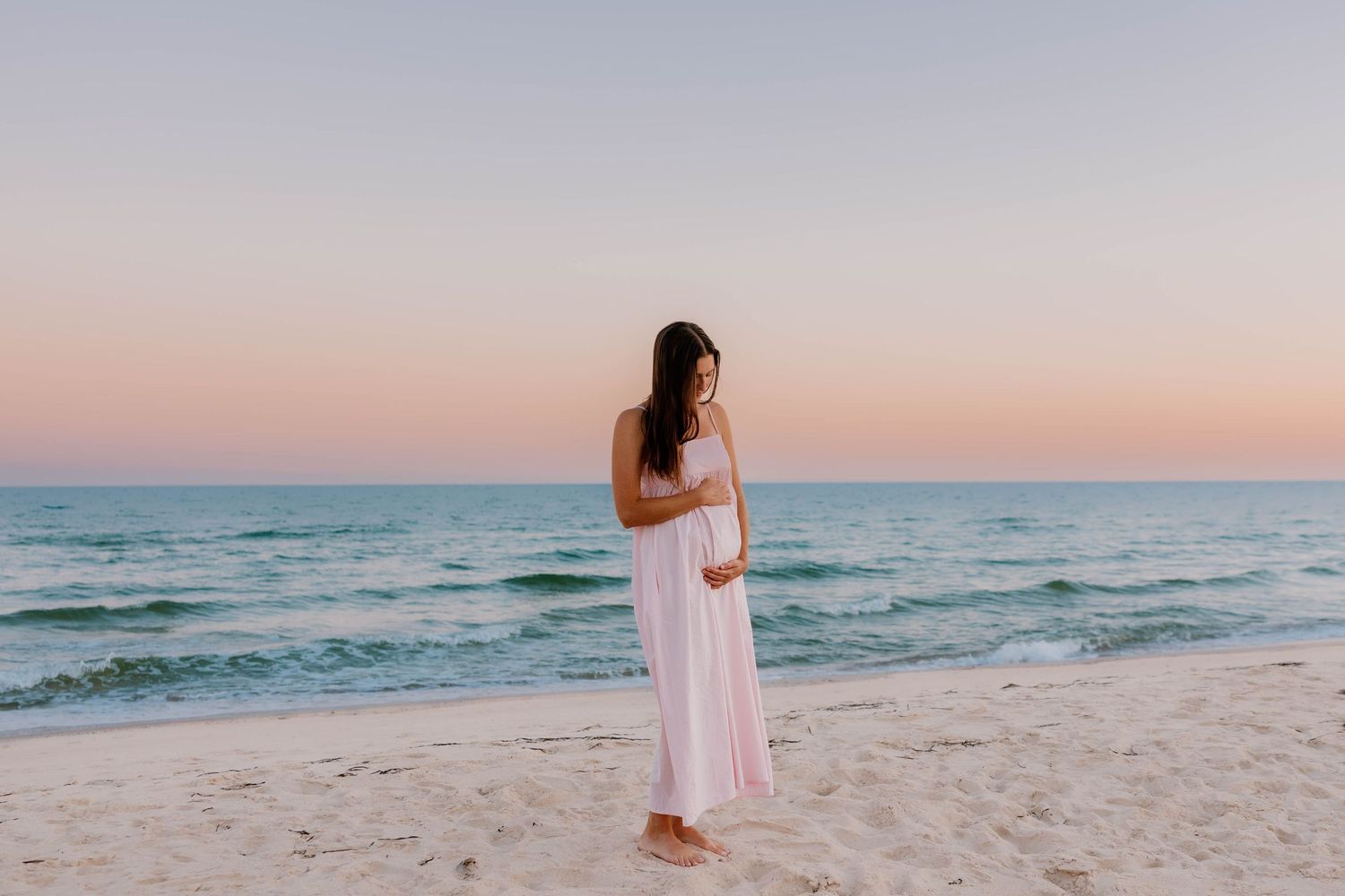 Pregnant woman in white maxi dress standing on sandy beach at sunset with turquoise ocean in background.