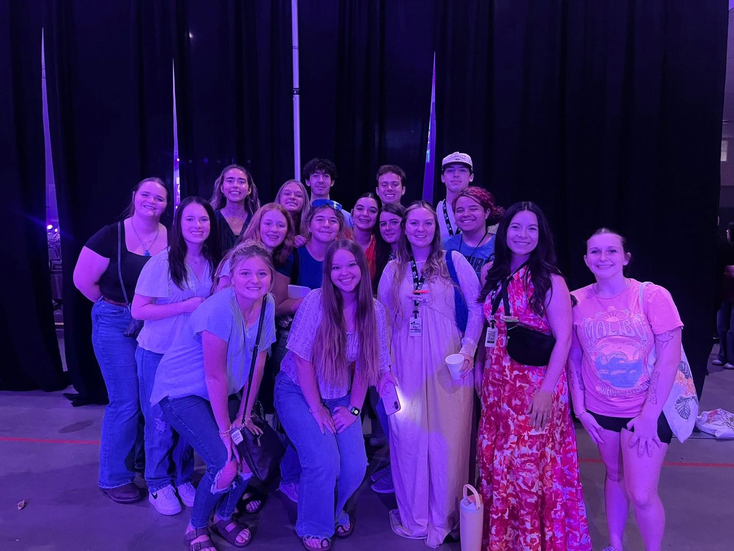 Large group of young adults posing together at a concert venue under purple stage lighting.