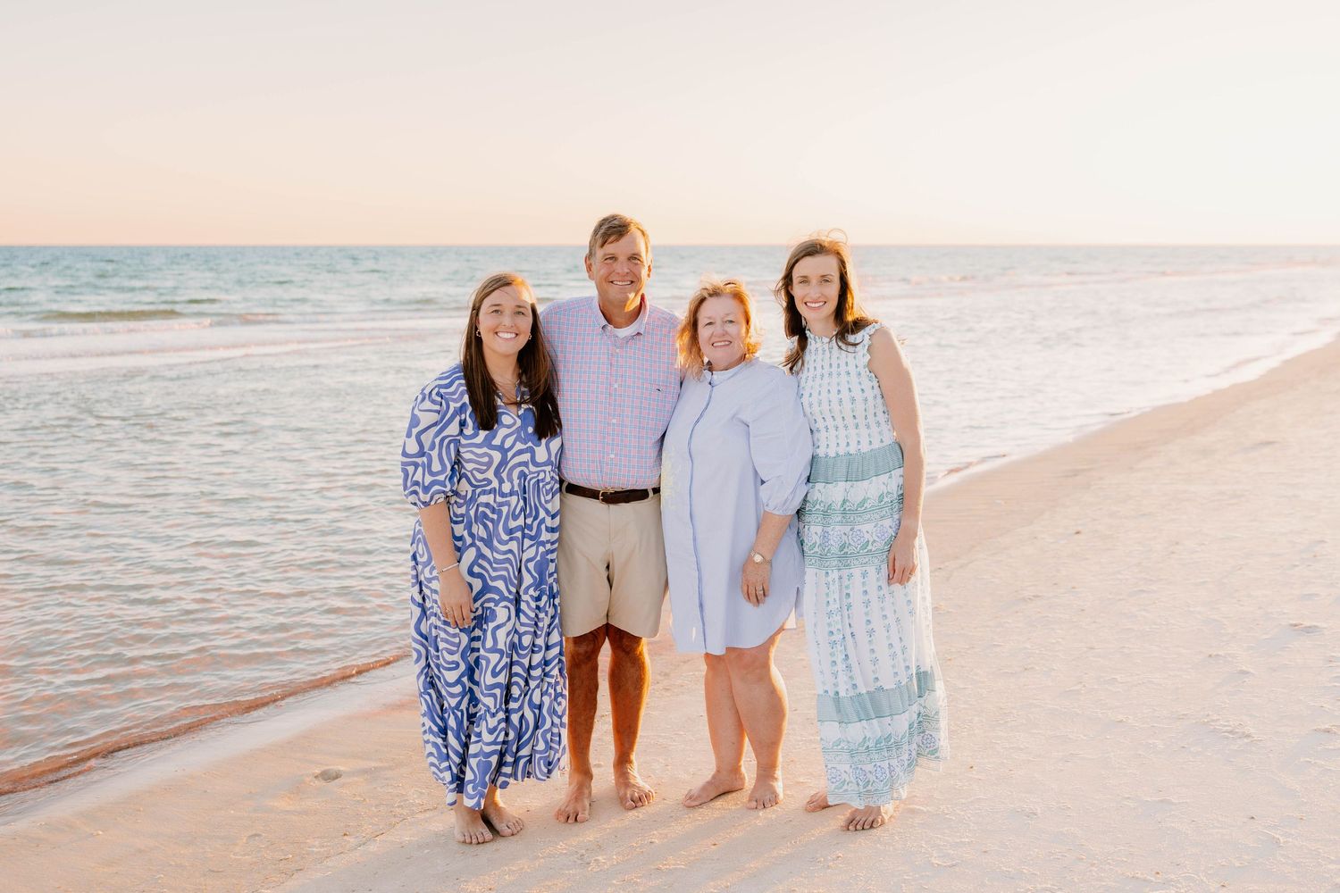 Family of four smiling together on a sandy beach at sunset, wearing casual summer outfits near calm ocean waters.