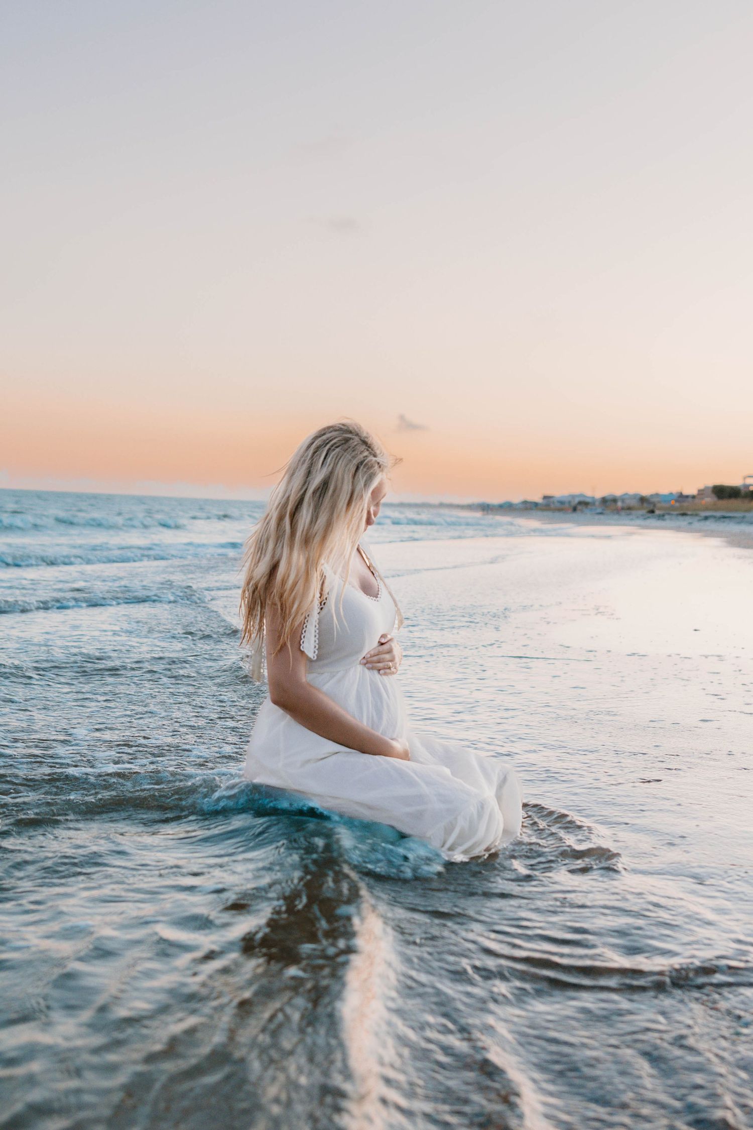 Pregnant woman in white dress sitting in ocean waves at sunset, cradling her baby bump on a serene beach.