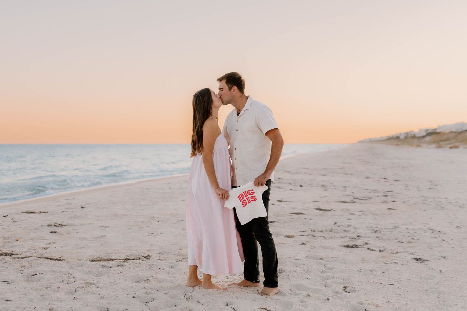 Couple kissing on a sandy beach at sunset, holding a baby announcement shirt in a romantic maternity reveal photo.