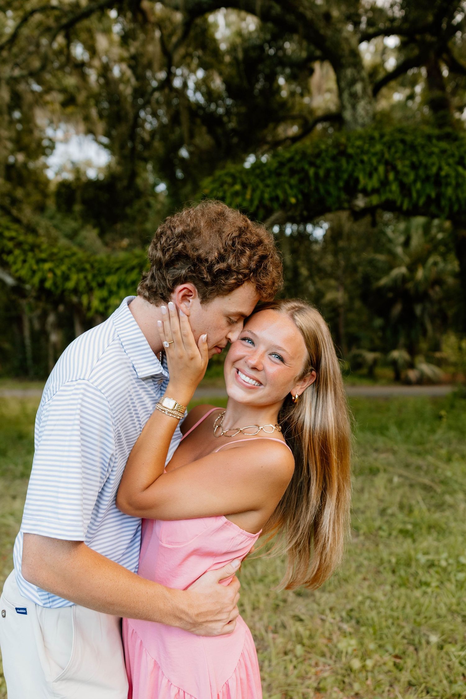 A couple embraces outdoors in a romantic moment, with the woman wearing a pink dress and the man in a striped shirt.