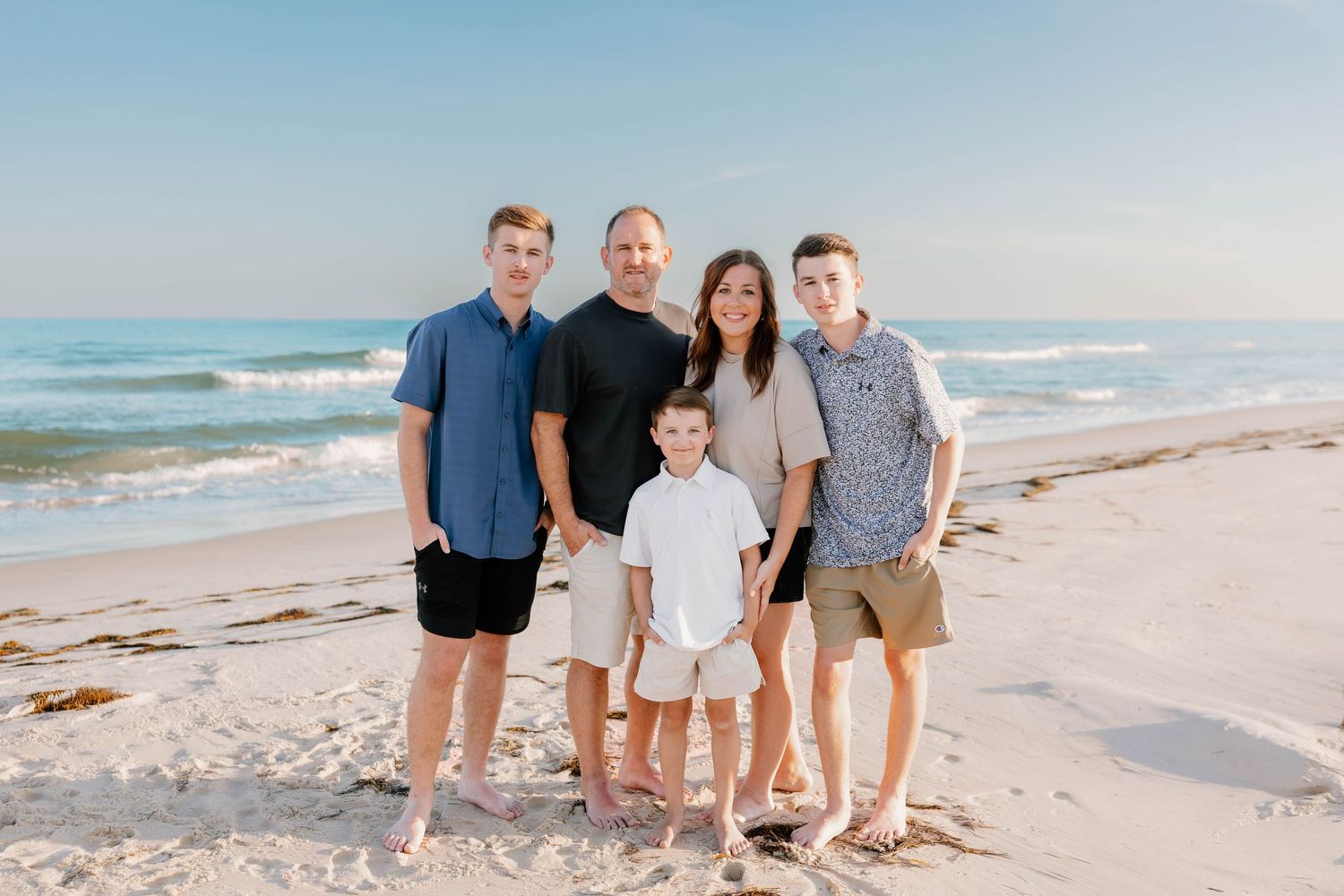 Family of five smiling together on a sunny beach with turquoise waves, wearing casual summer outfits.