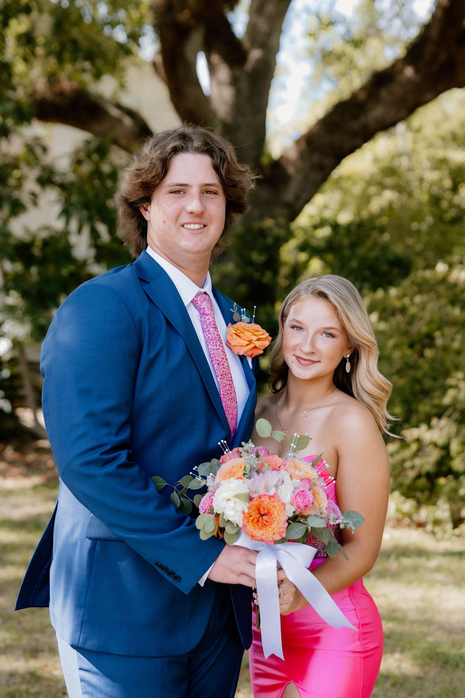 A couple dressed for prom, man in blue suit with boutonniere, woman in pink dress holding colorful bouquet.