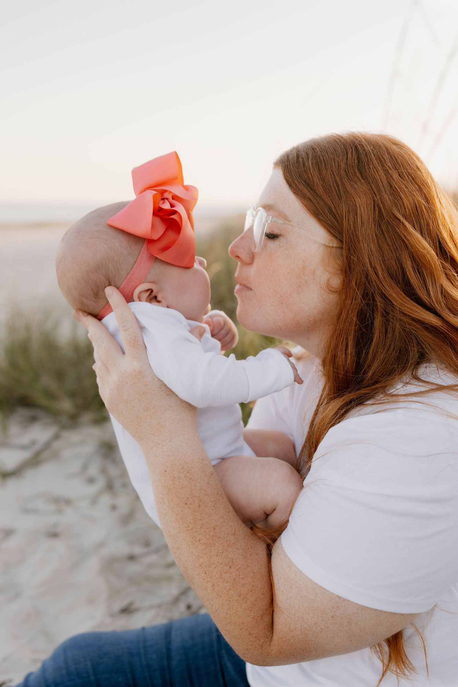 A freckled redhead mother kisses her baby girl wearing a coral bow headband on a sandy beach.
