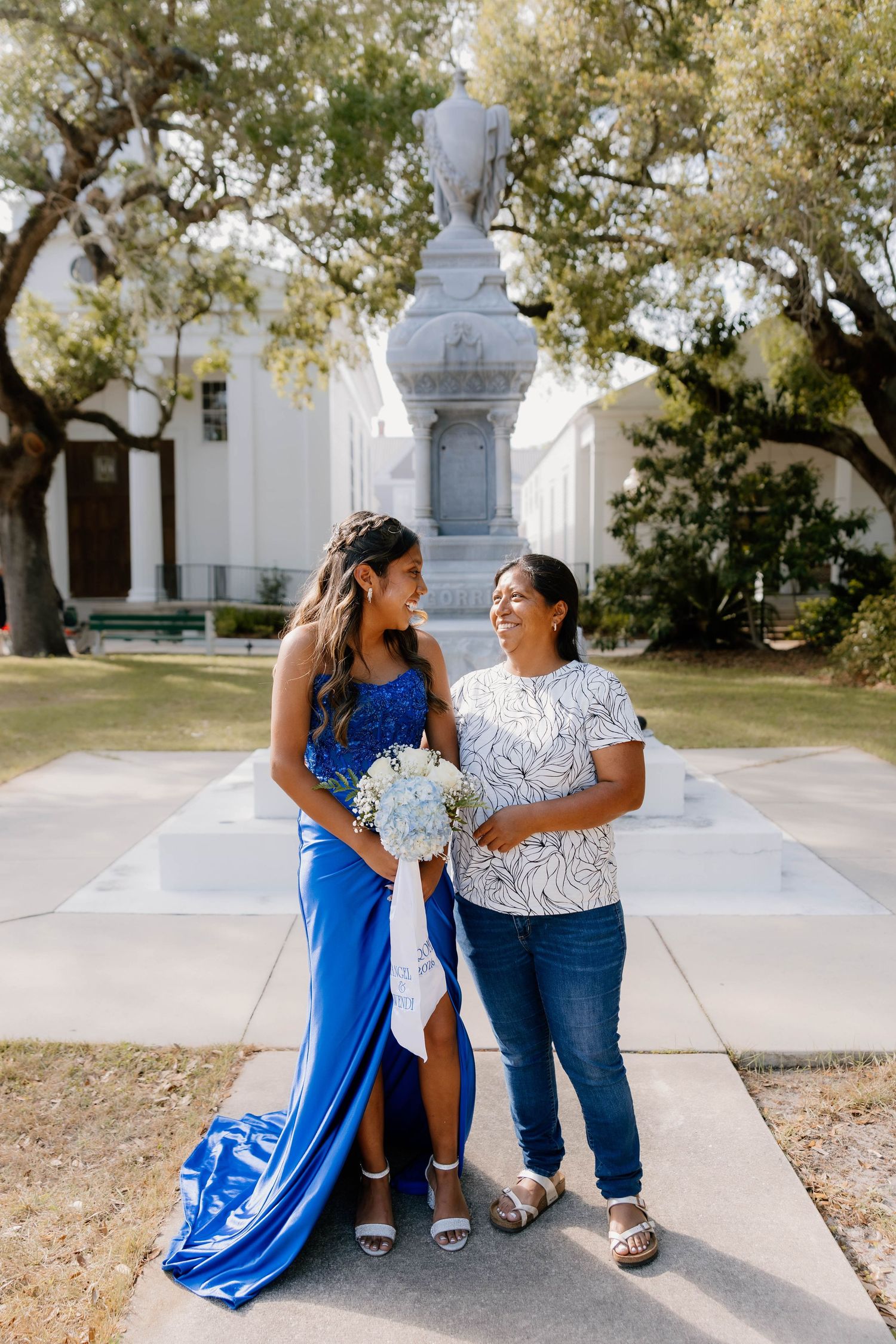 Young woman in royal blue gown holding bouquet poses with older woman in floral top outdoors near monument.