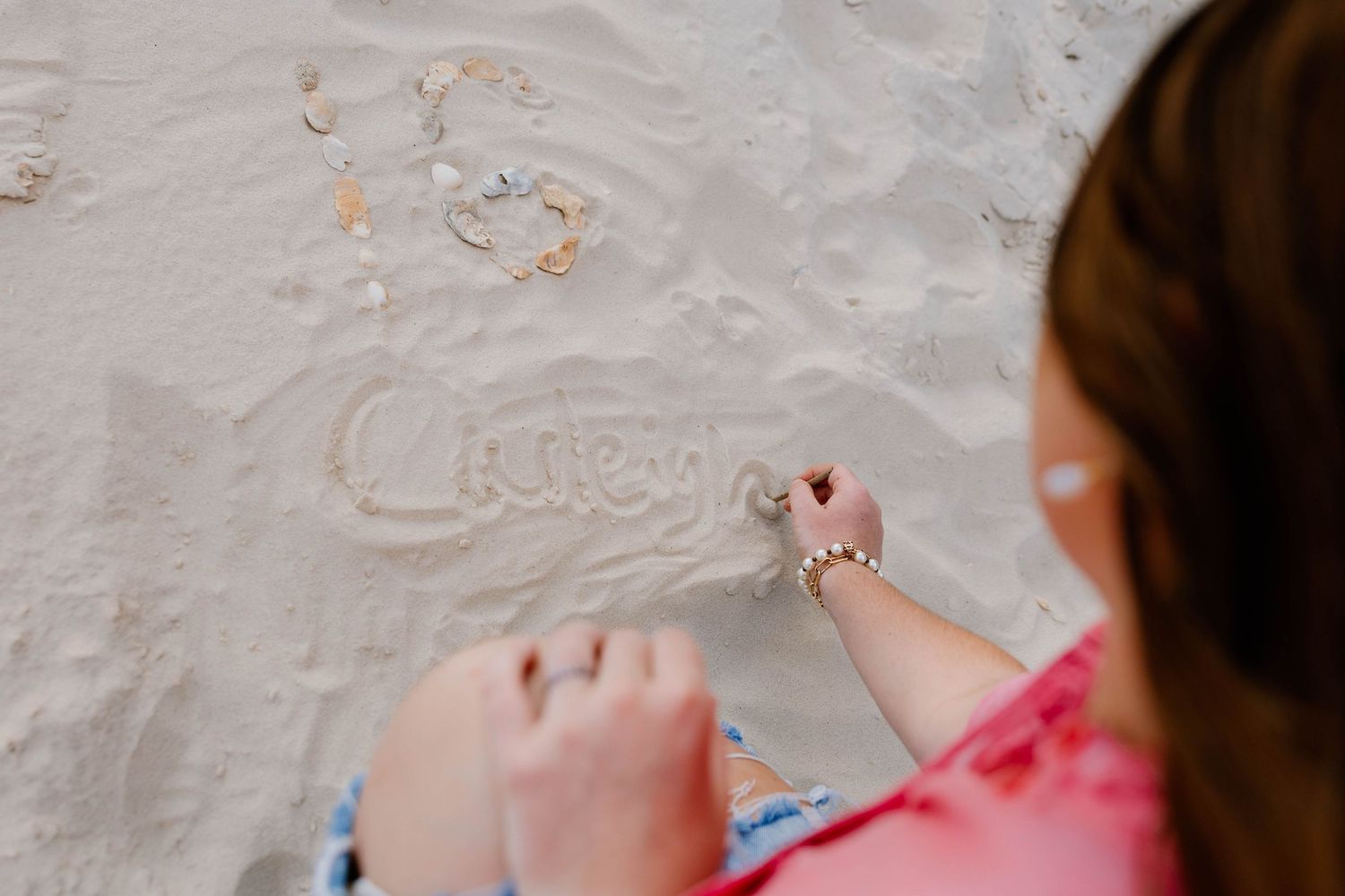 A woman writes a name in the sand on a sunny beach, wearing a pink top and a beaded bracelet.