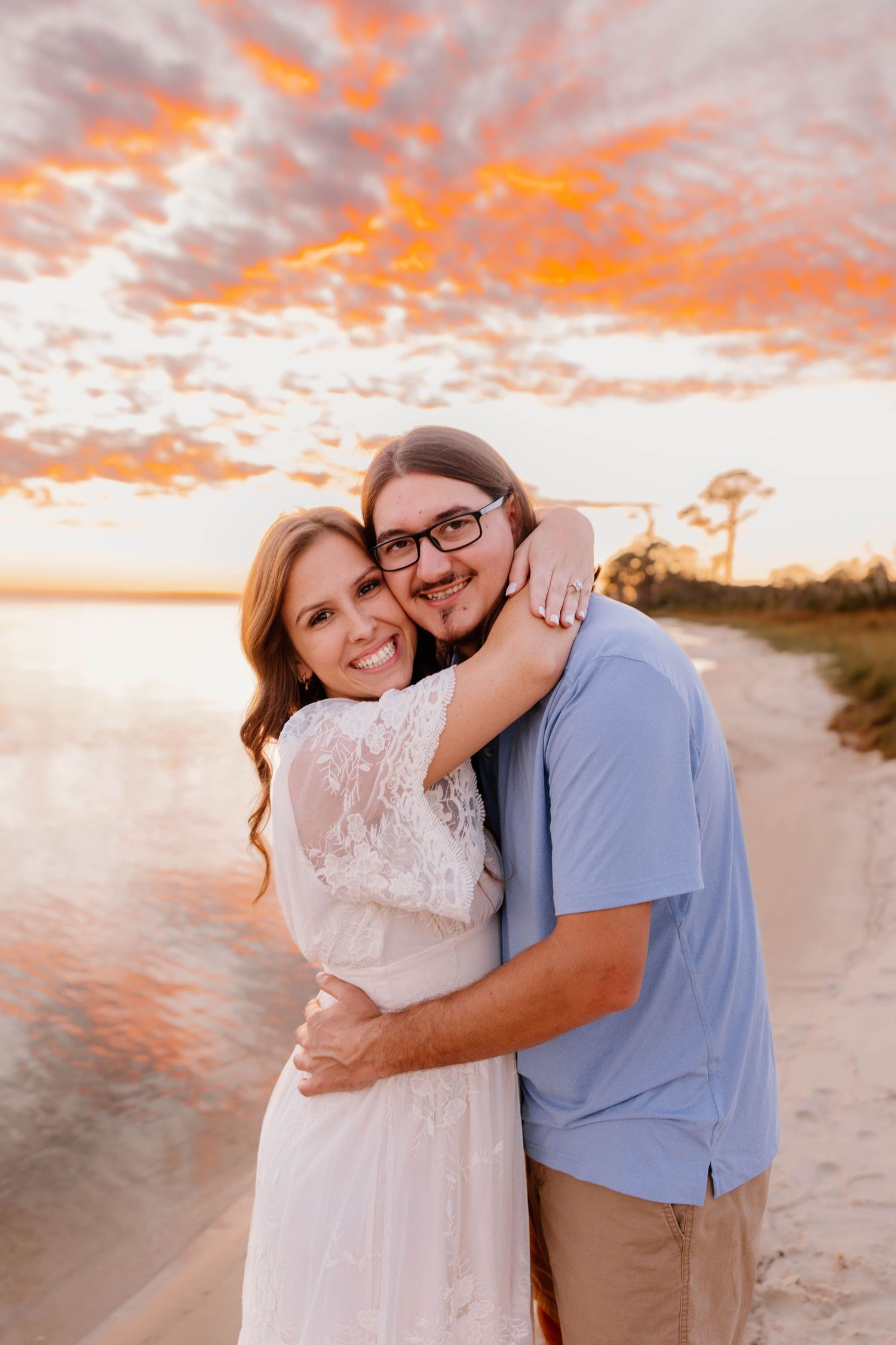 Couple embracing on beach at sunset with vibrant orange sky, woman in white lace dress and man in blue shirt smiling.