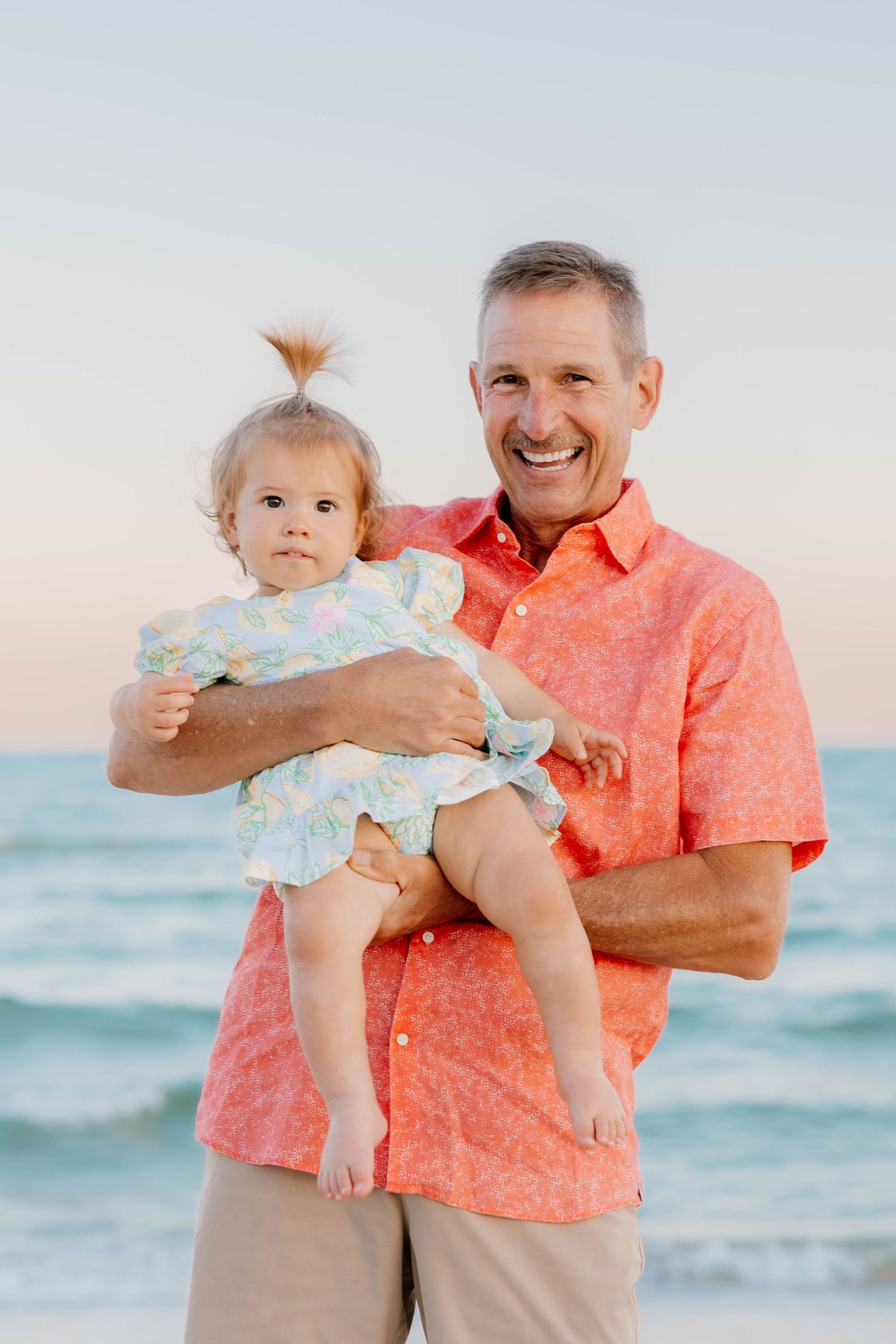 A loving moment shared between a senior in coral shirt and young child against a serene beach backdrop at sunset.