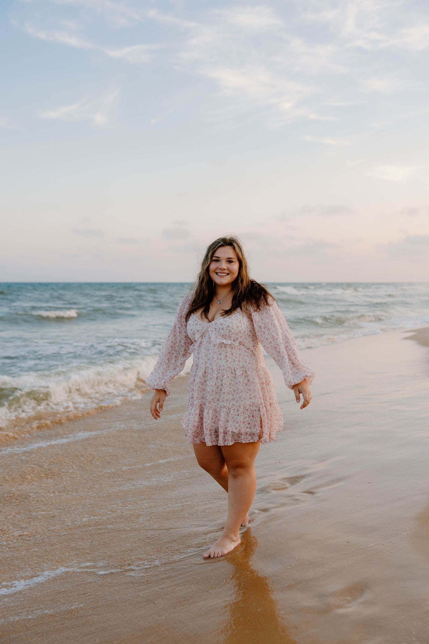 Young woman in floral mini dress walking barefoot on sandy beach with ocean waves at sunset.