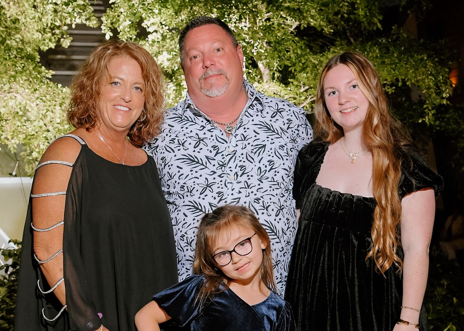 Family of four posing together at an elegant outdoor evening event, with lush greenery in the background.