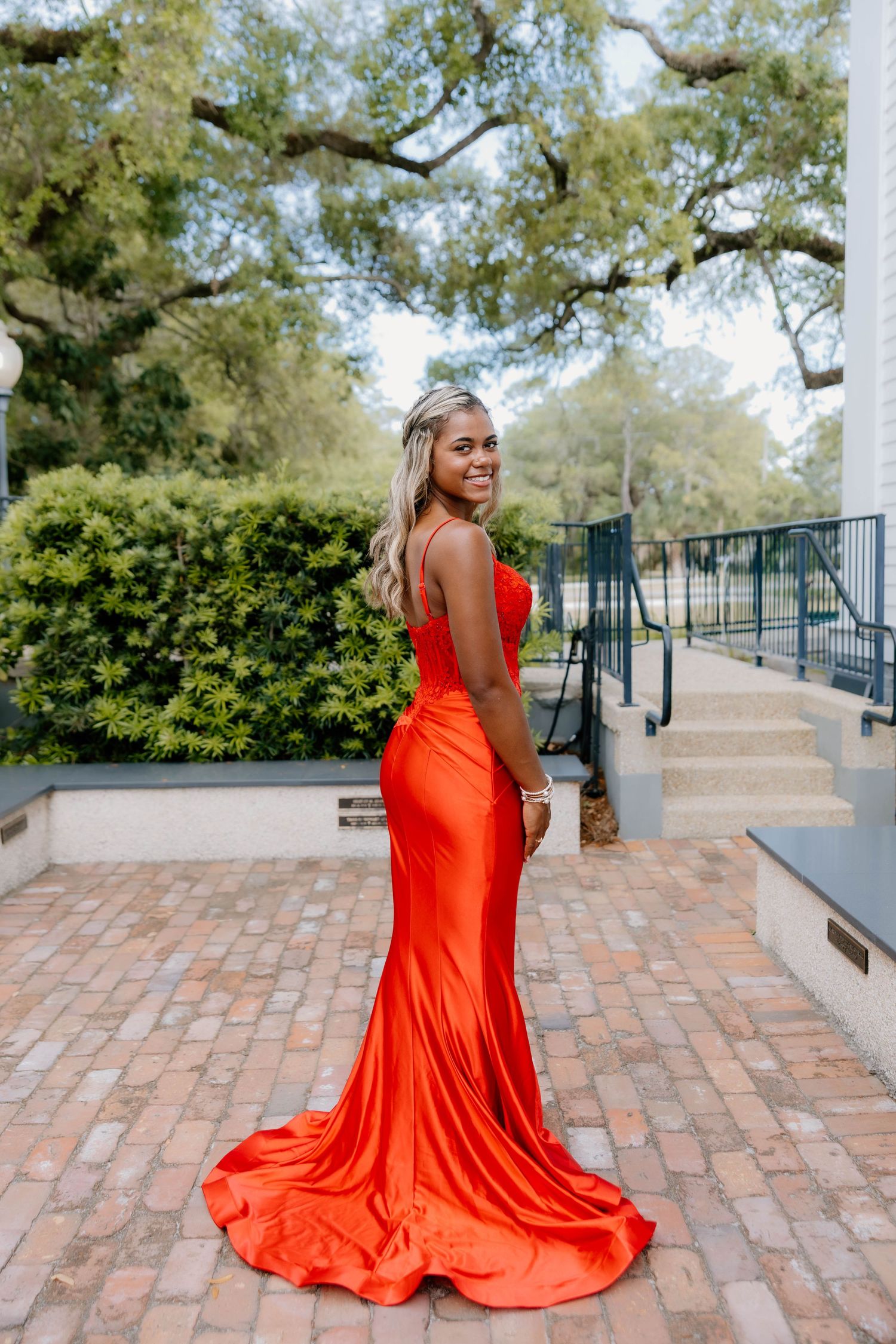 Young woman in elegant red satin mermaid gown posing outdoors on brick pathway with lush greenery backdrop.