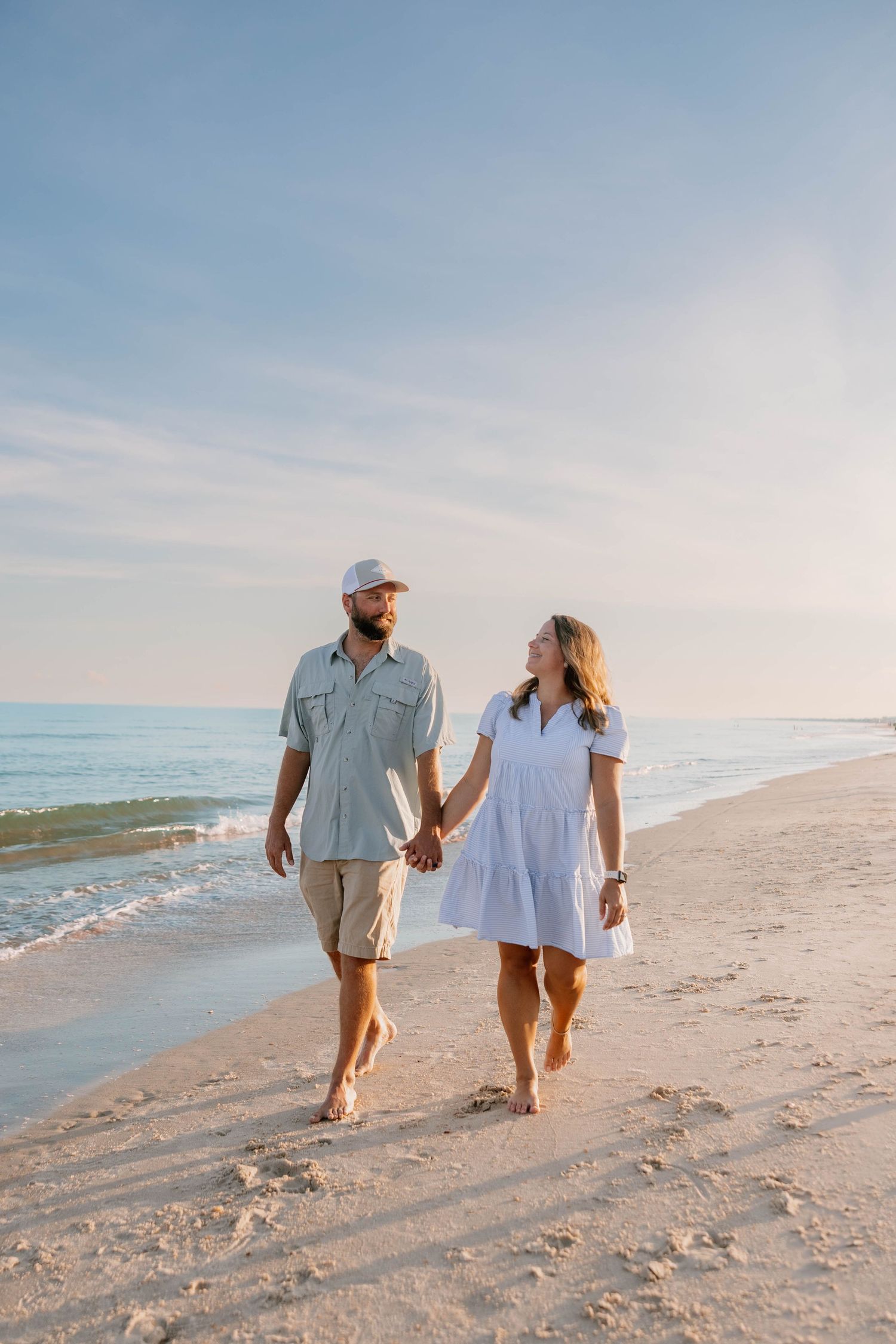 Couple holds hands while walking barefoot on beach at sunset.