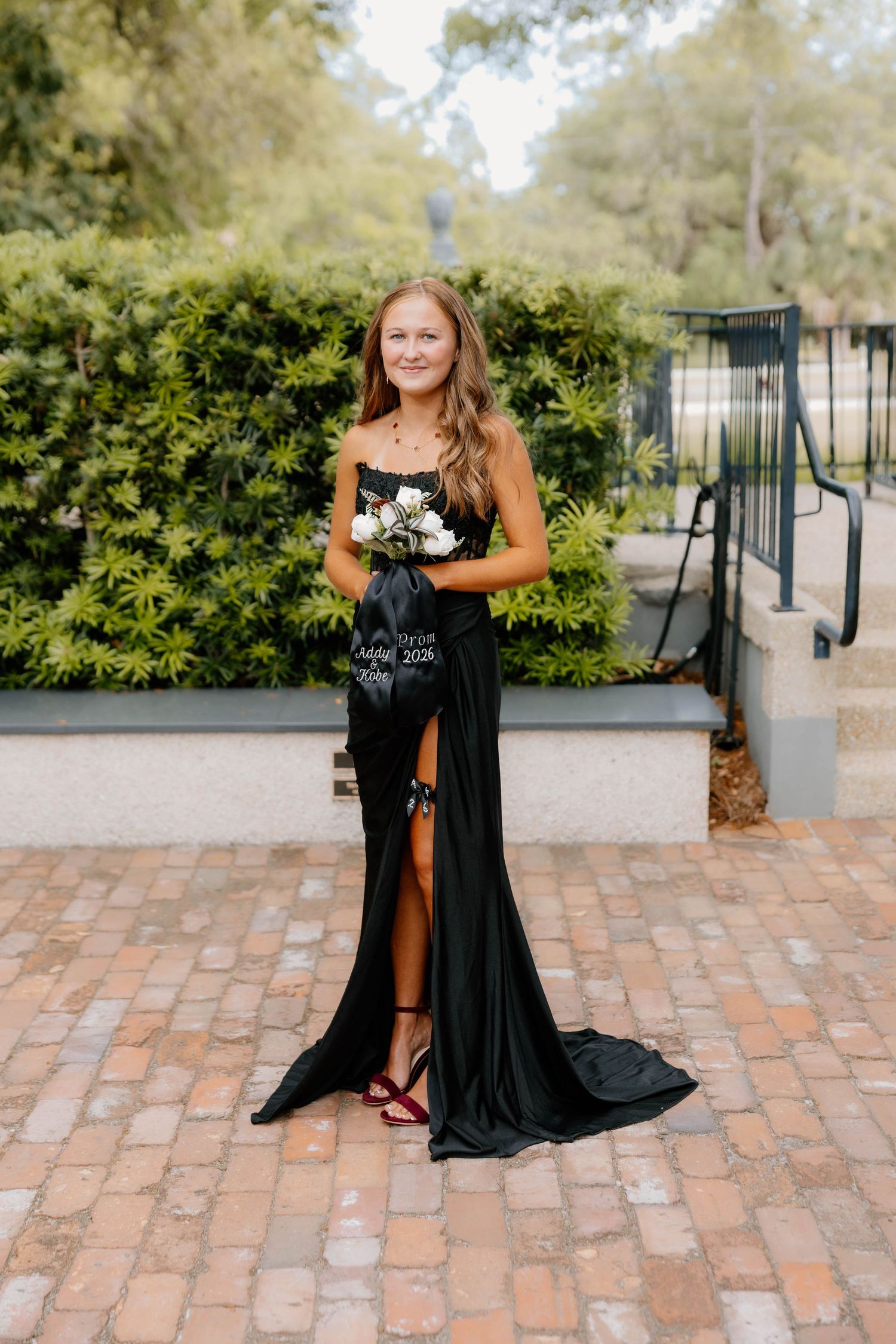 Young woman in elegant black satin gown holding white flowers, posing on brick pathway at formal event.