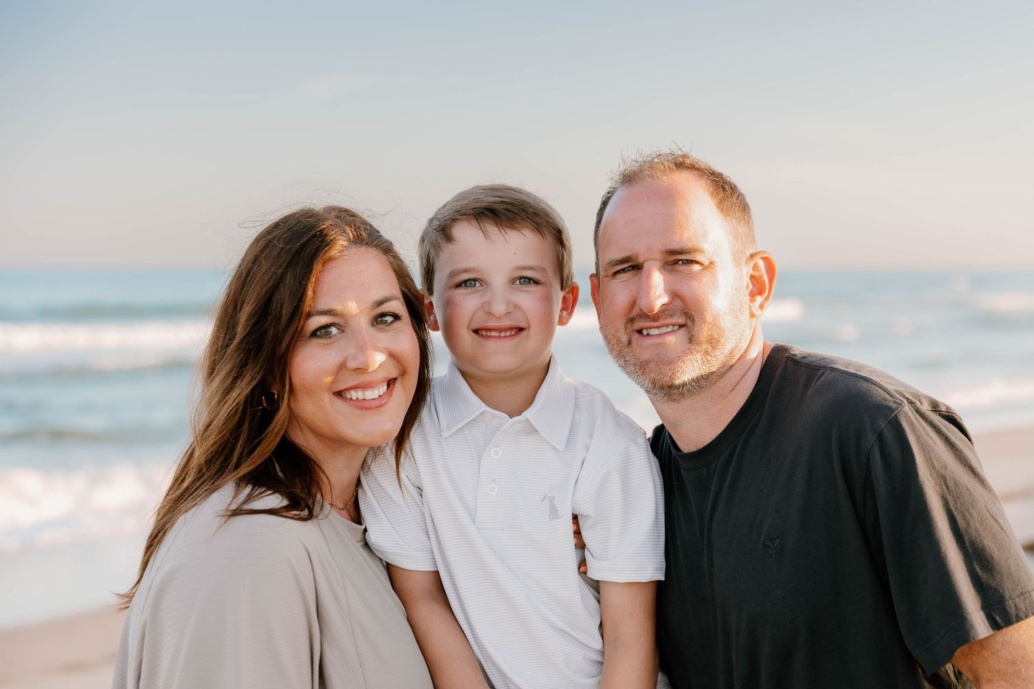 Family of three smiling together on a sunny beach, with ocean waves in the background.