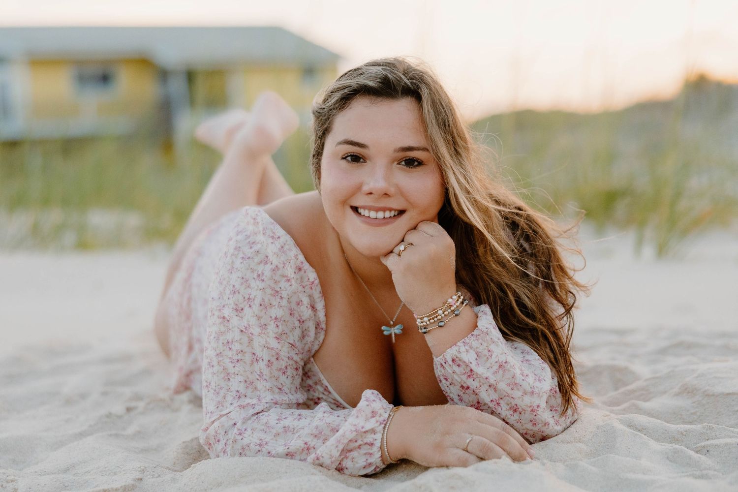 Smiling teenage girl lying on sandy beach in floral dress, golden hour light, with colorful house in background.