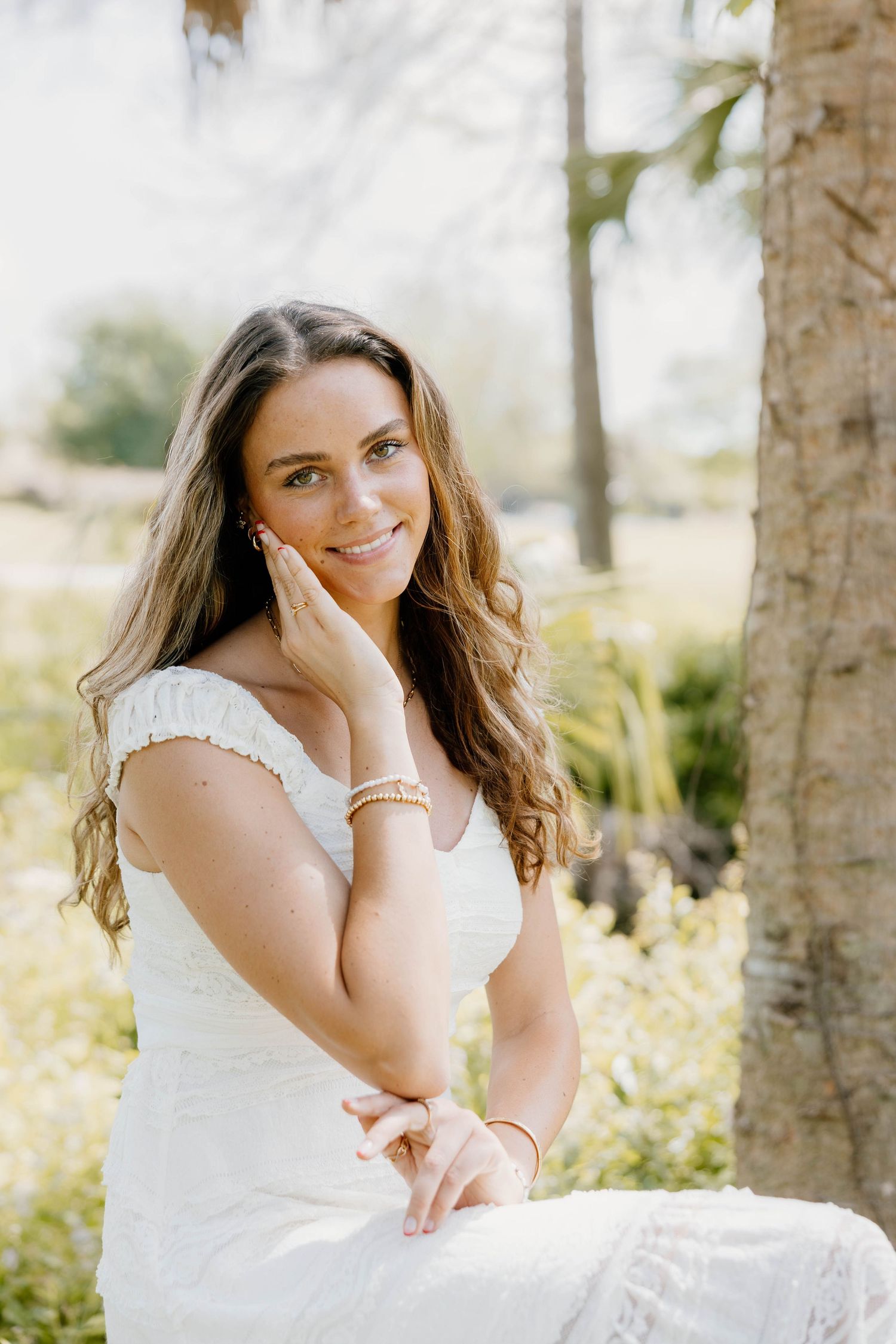 Young woman in white lace dress smiling outdoors, posing gracefully near a tree in a sunlit garden.