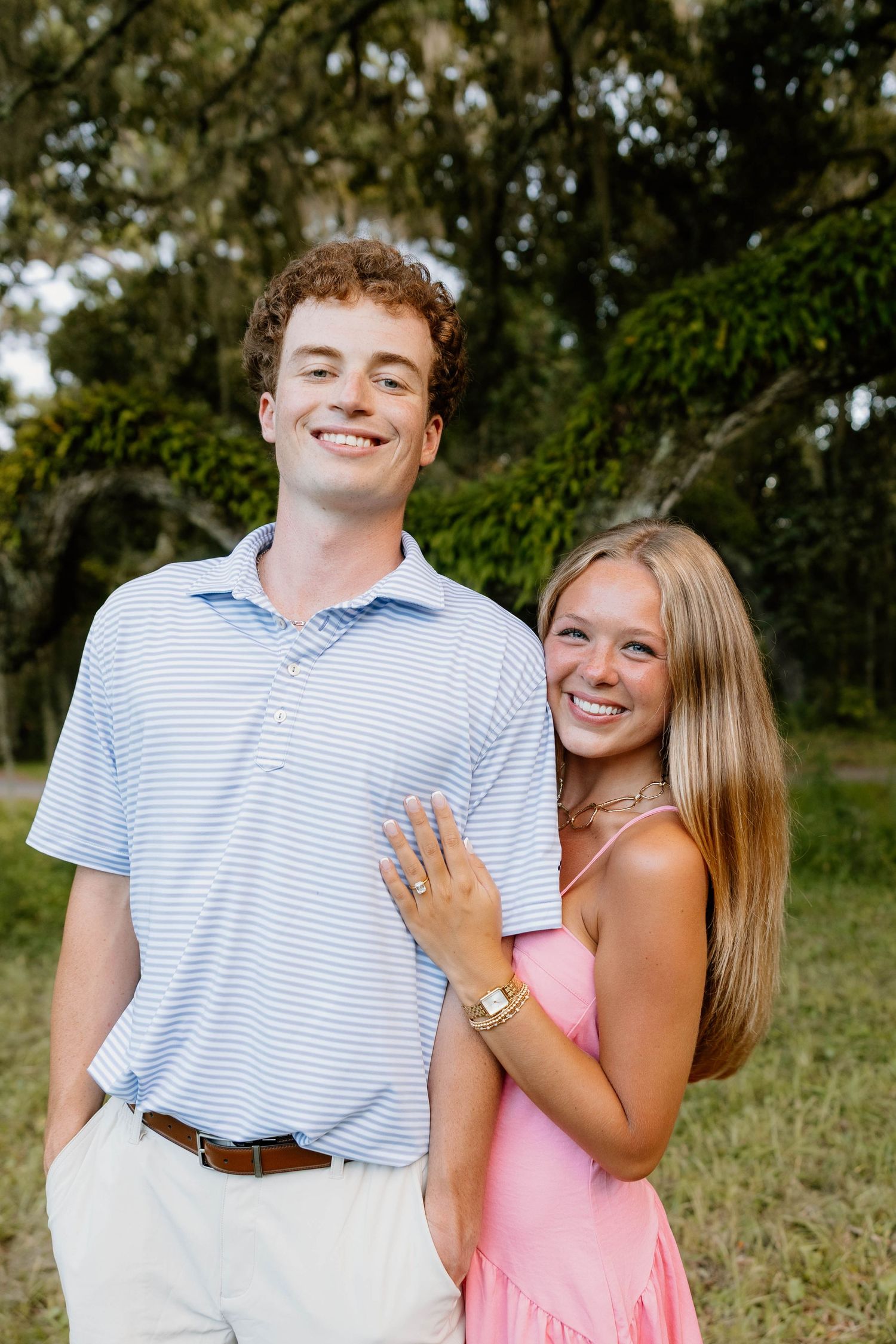 A smiling young couple poses outdoors, man in striped polo and woman in pink dress, surrounded by lush green trees.