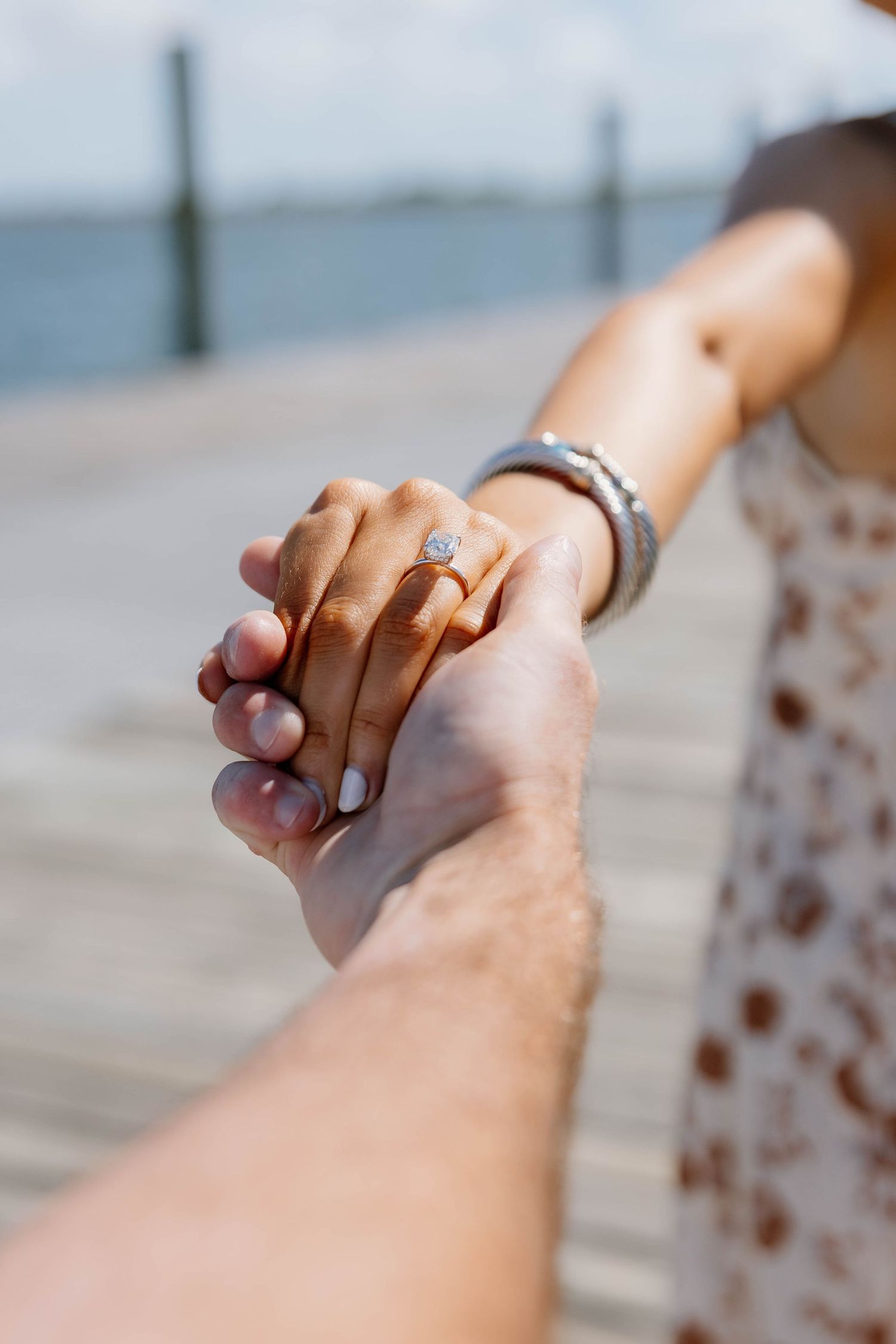 Close-up of a woman's hand displaying a diamond engagement ring while holding her partner's hand on a dock.