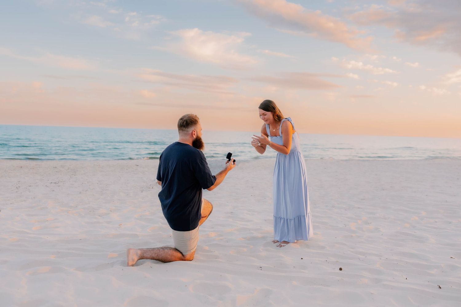 A couple in formal attire poses for romantic photos on a white sandy beach at sunset.