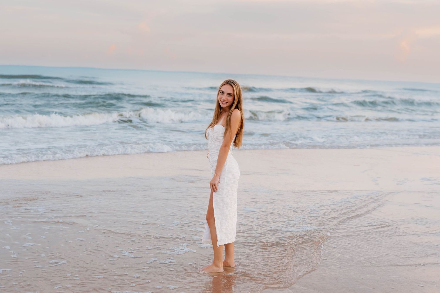 A young woman in a white midi dress stands barefoot on a sandy beach with ocean waves in the background.