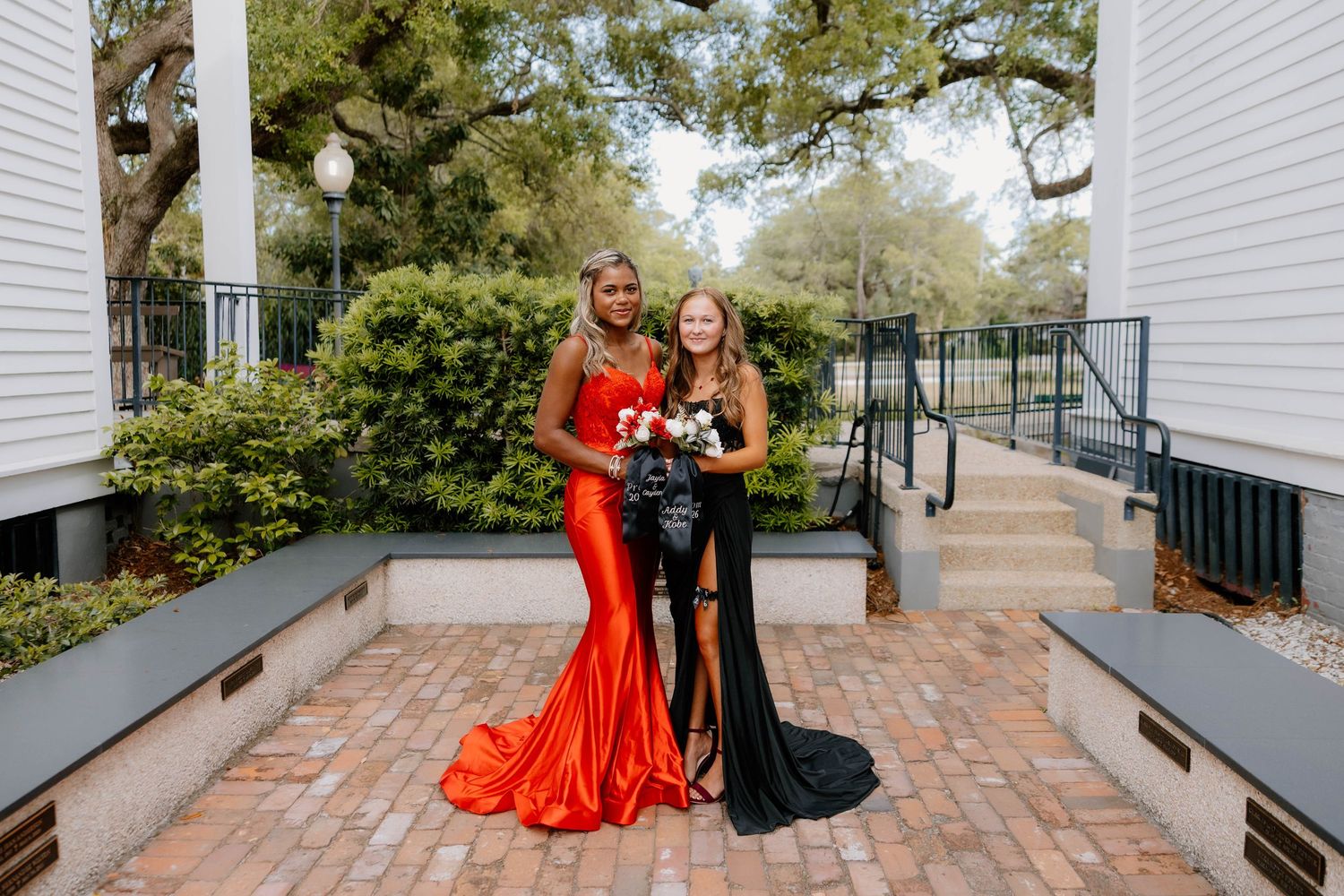 Two prom girls pose outdoors, one in red gown and one in black sequin dress, holding bouquets.