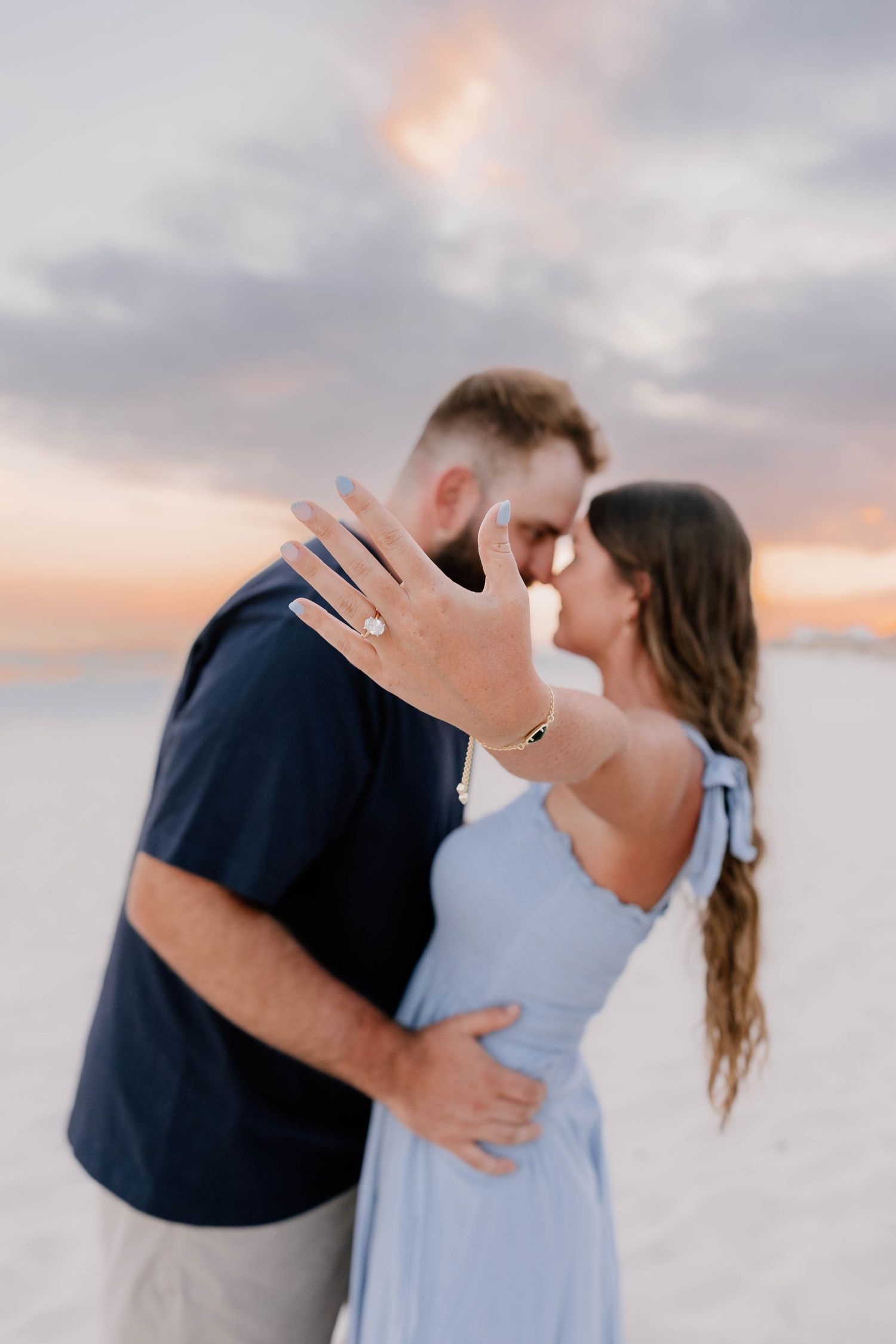 A couple embraces romantically on a beach at sunset with soft pink and purple clouds in the background.