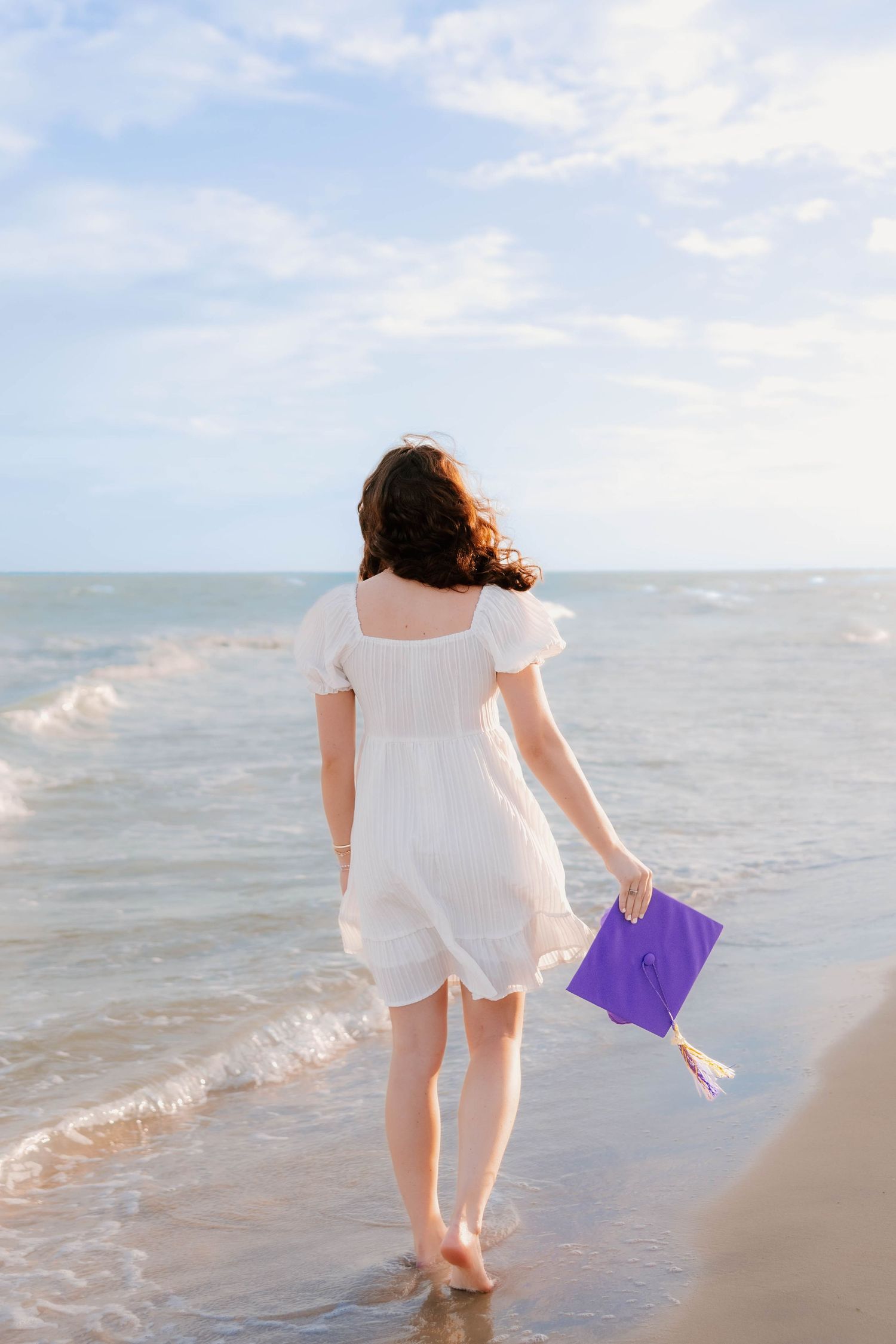 A young woman in a white dress holds a purple graduation cap while standing barefoot on a sandy beach shoreline.