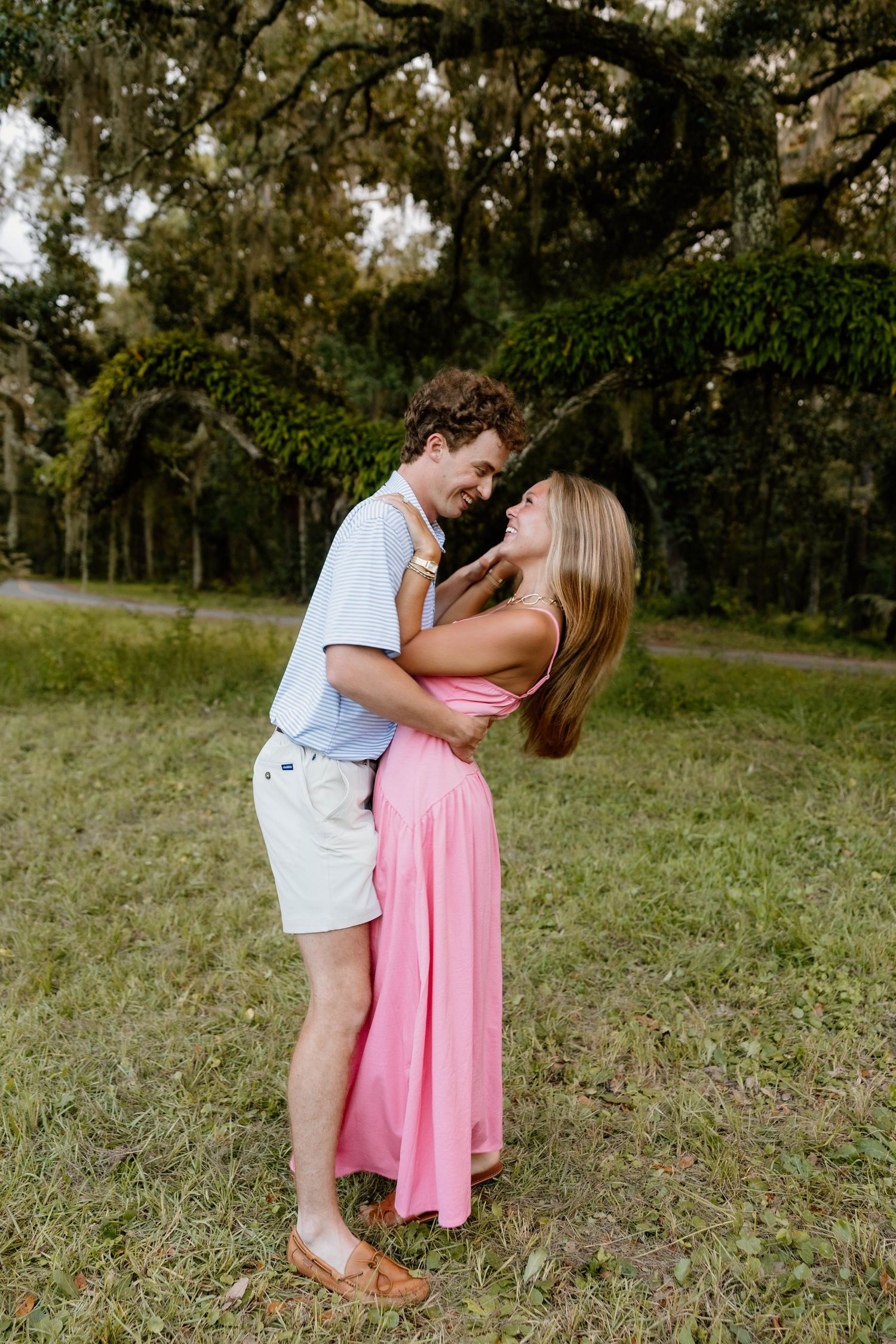 A couple embraces and gazes at each other lovingly in a lush green park, woman wearing a pink maxi dress.