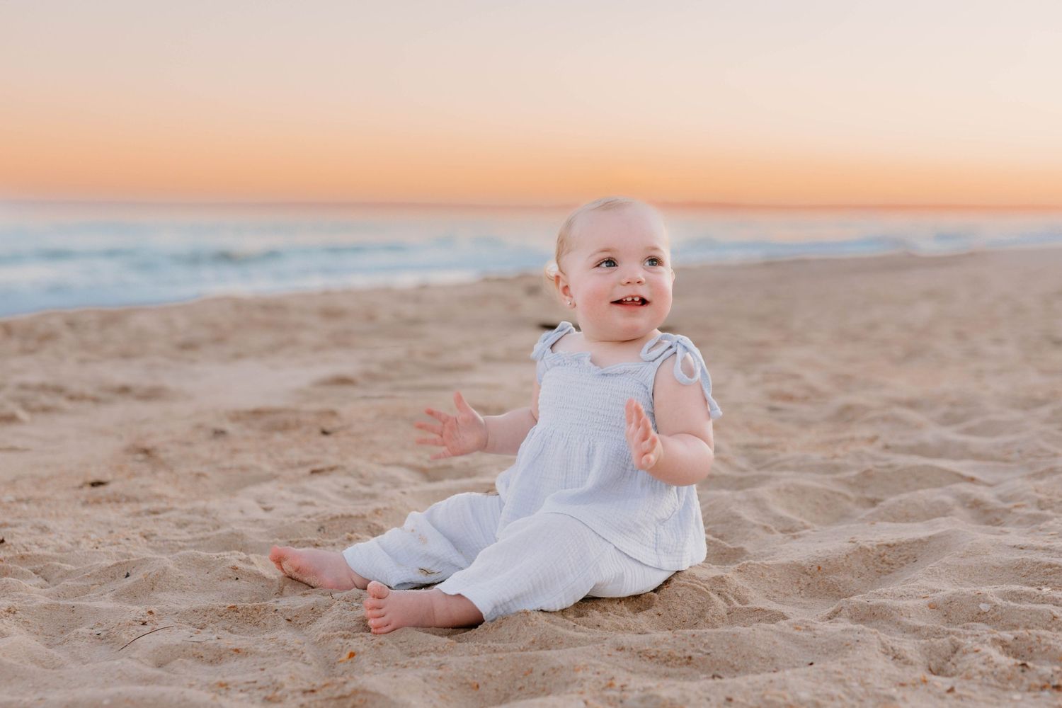 Happy baby in white outfit sitting on sandy beach at sunset with ocean waves in background.