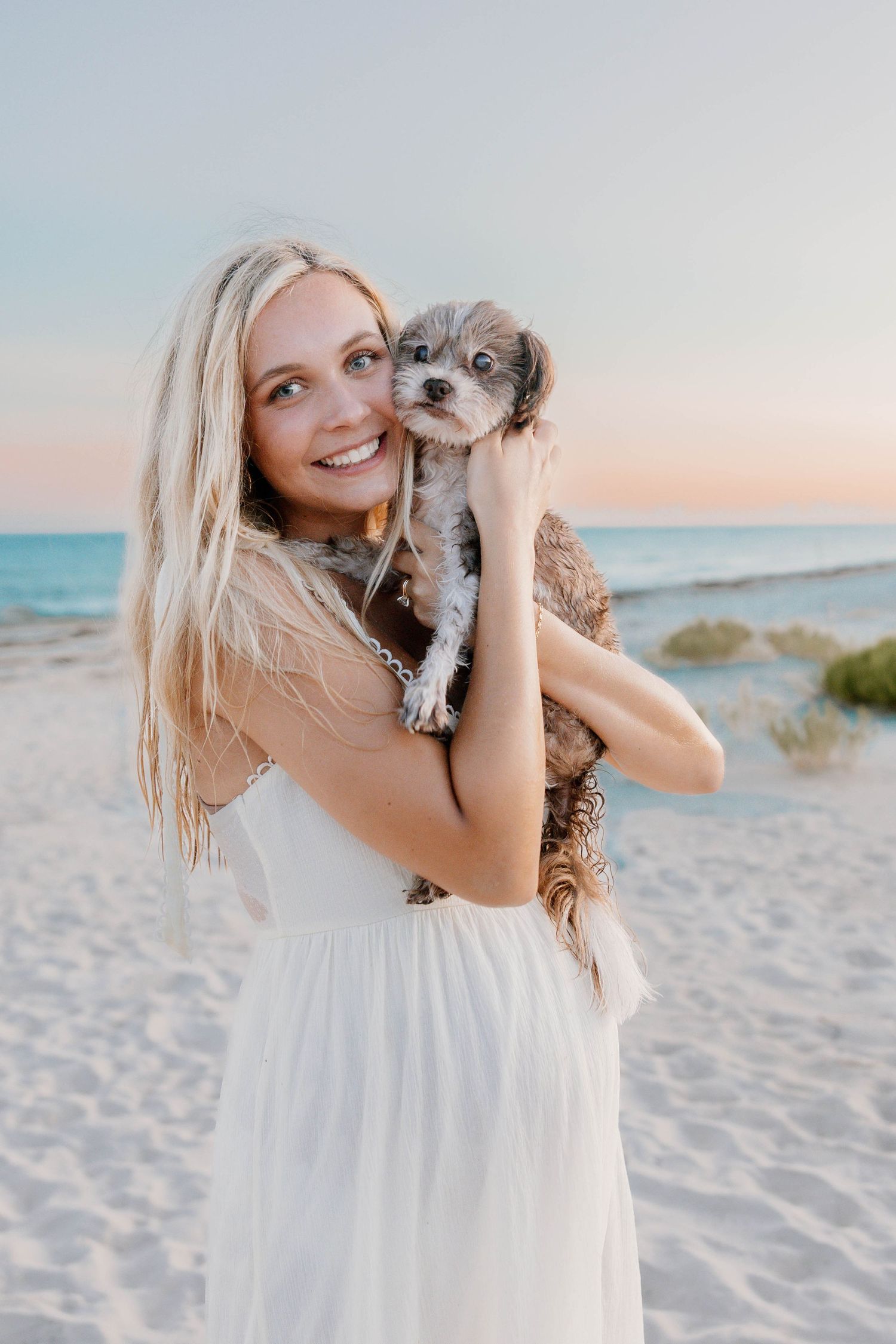 A person in a white dress holds and plays with a small dog on a sunny beach at sunset.
