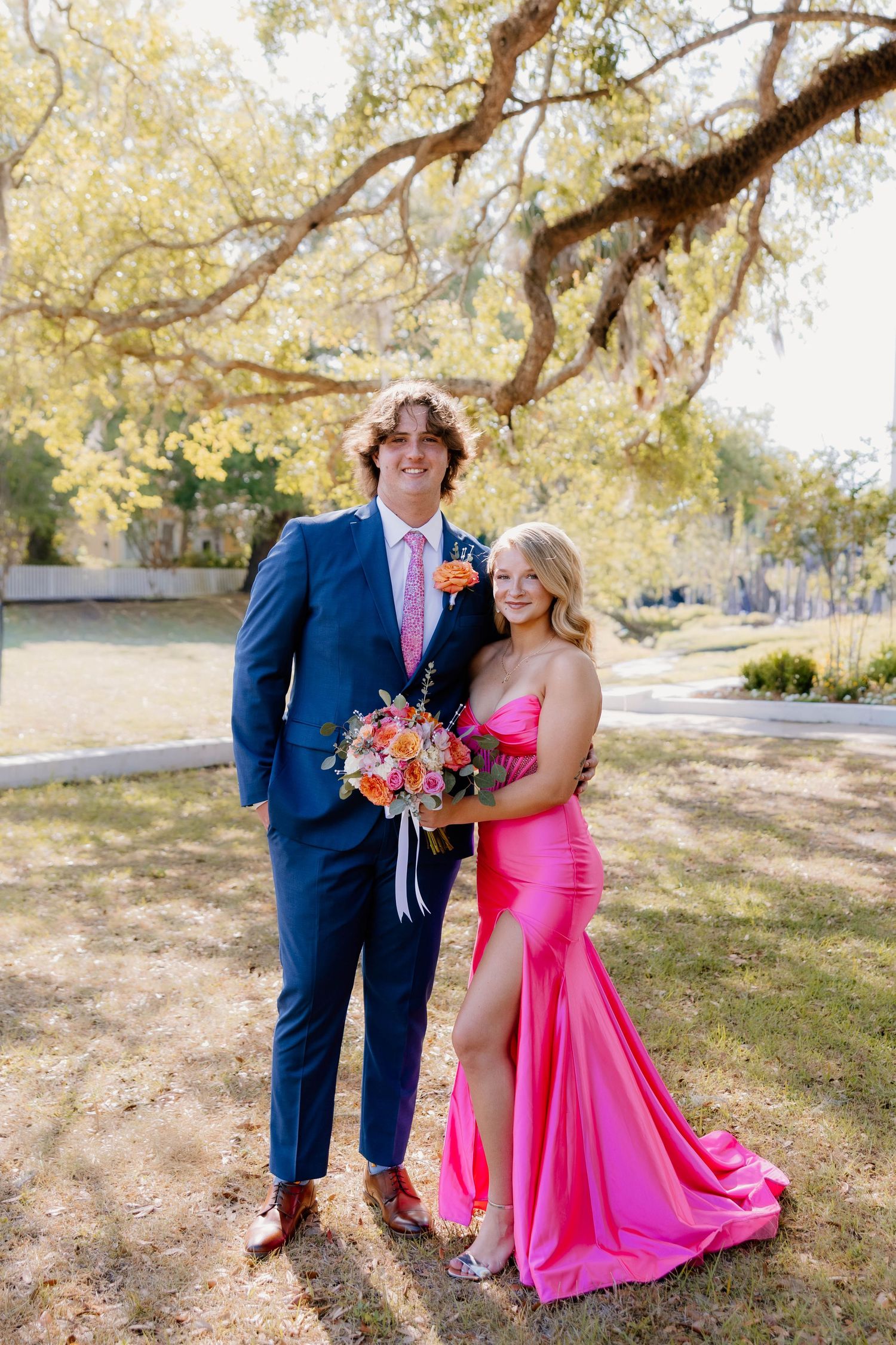 A young couple dressed for prom, him in a navy suit with pink tie, her in a hot pink gown, holding a colorful bouquet outdoors.