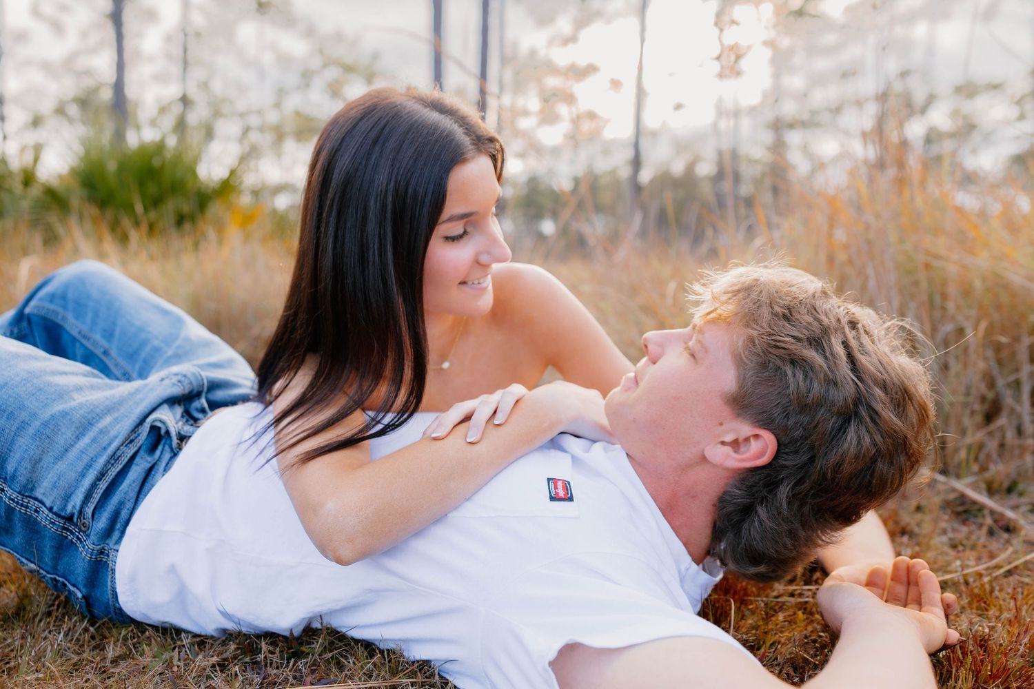 Couple lying in golden grass field, woman in jeans smiling at man in white shirt during outdoor engagement session.