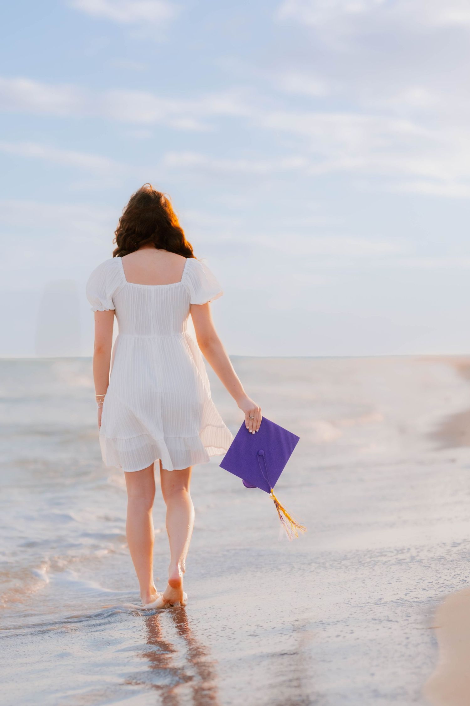 Young woman in white dress holding purple graduation cap walks barefoot along ocean shoreline at sunset.