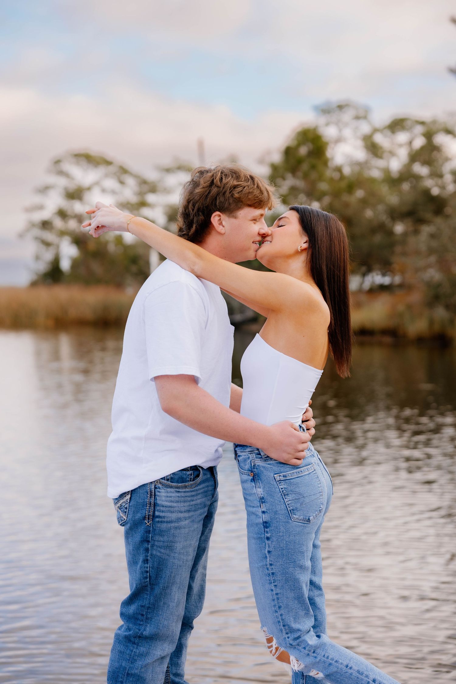 A couple sharing a romantic kiss by a serene lake, dressed in white tops and blue jeans, with trees and cloudy sky behind.