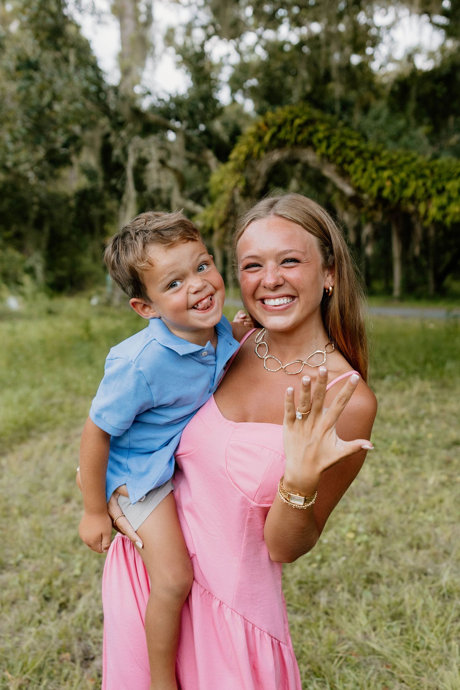 Smiling woman in pink dress holding a young boy while showing off her engagement ring outdoors.