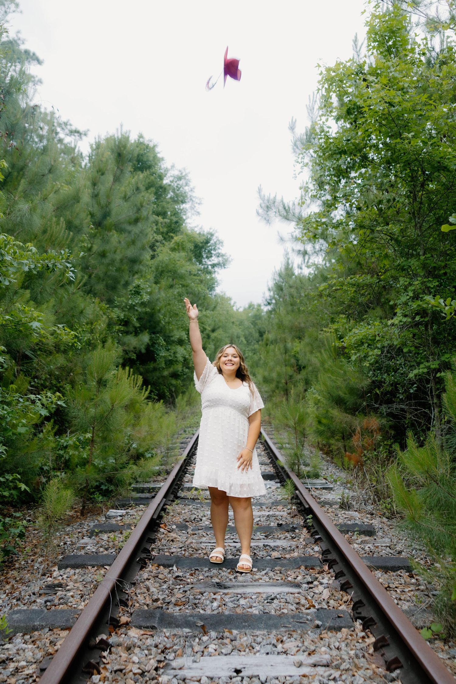 Woman in white dress standing on railroad tracks tossing a graduation cap into the air surrounded by lush green trees.