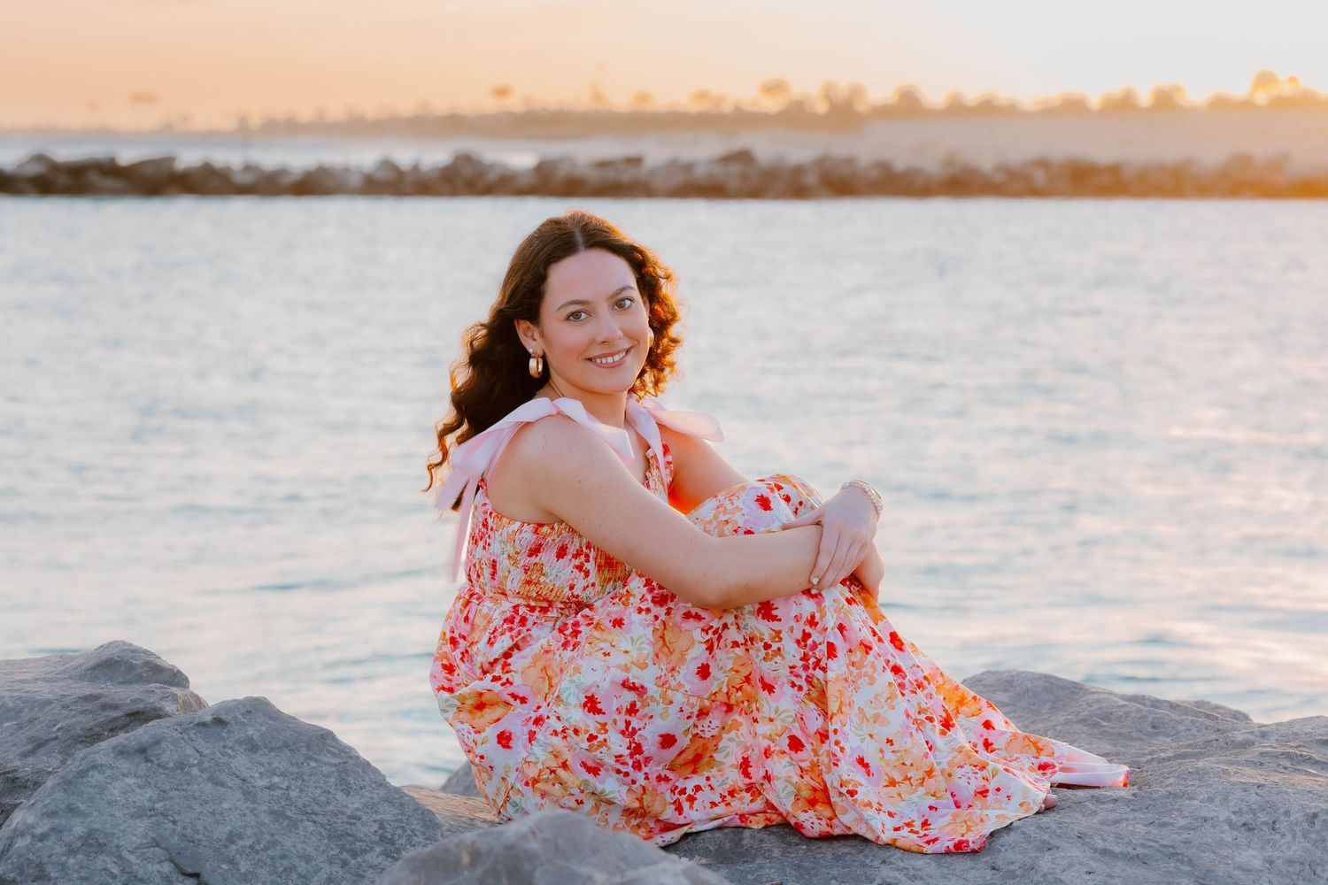 Woman in floral dress sitting on rocks by the ocean at golden hour sunset.
