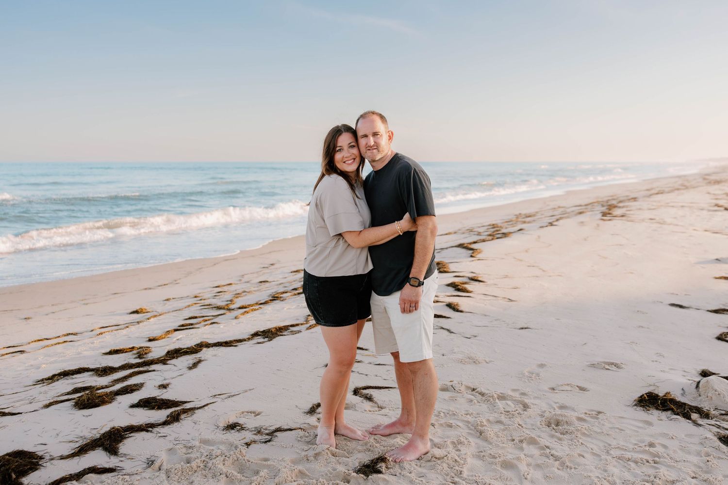 Couple embracing and smiling on a sandy beach at sunset with gentle ocean waves in the background.