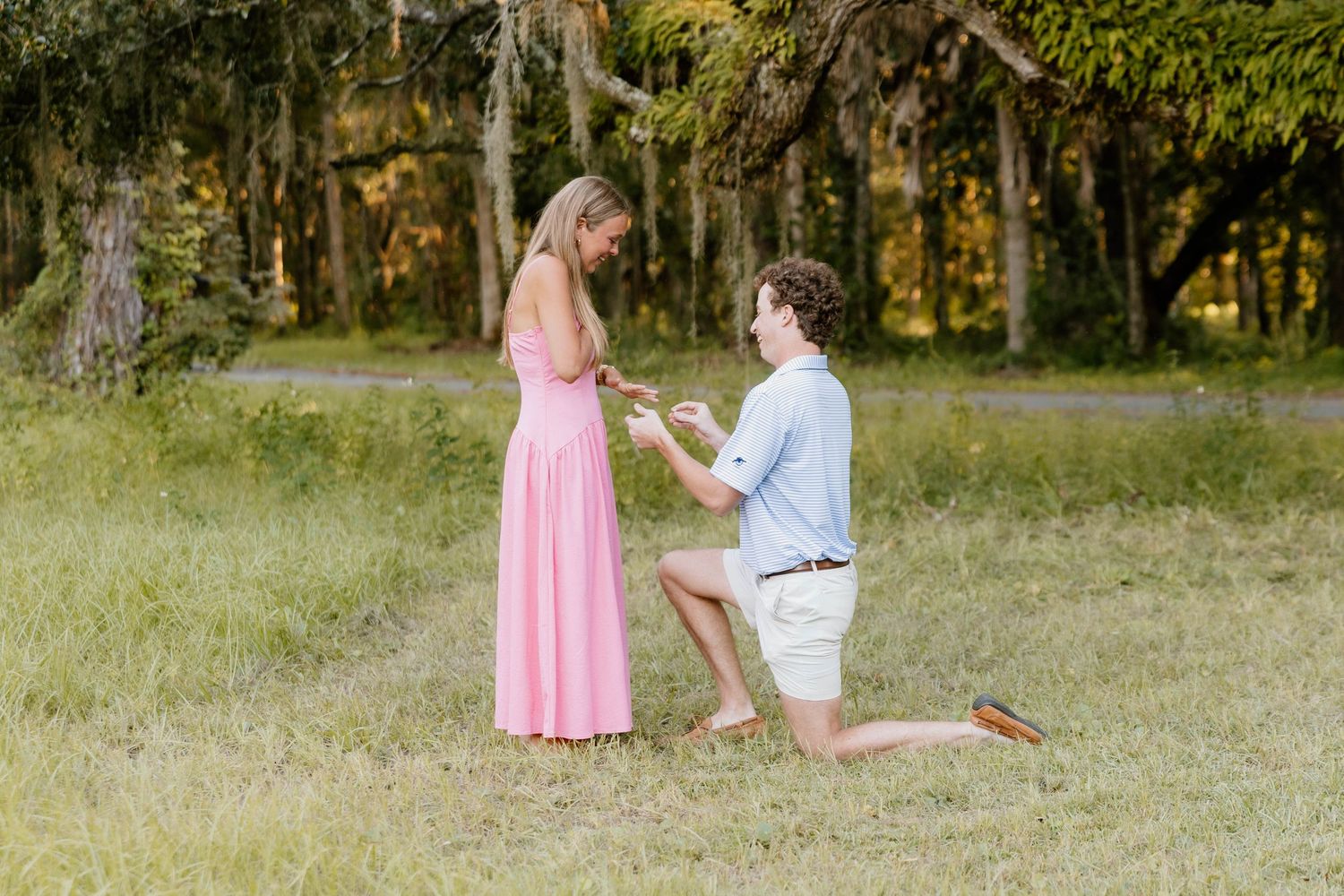 Man kneeling on one knee proposing to woman in pink dress outdoors in lush green park setting.