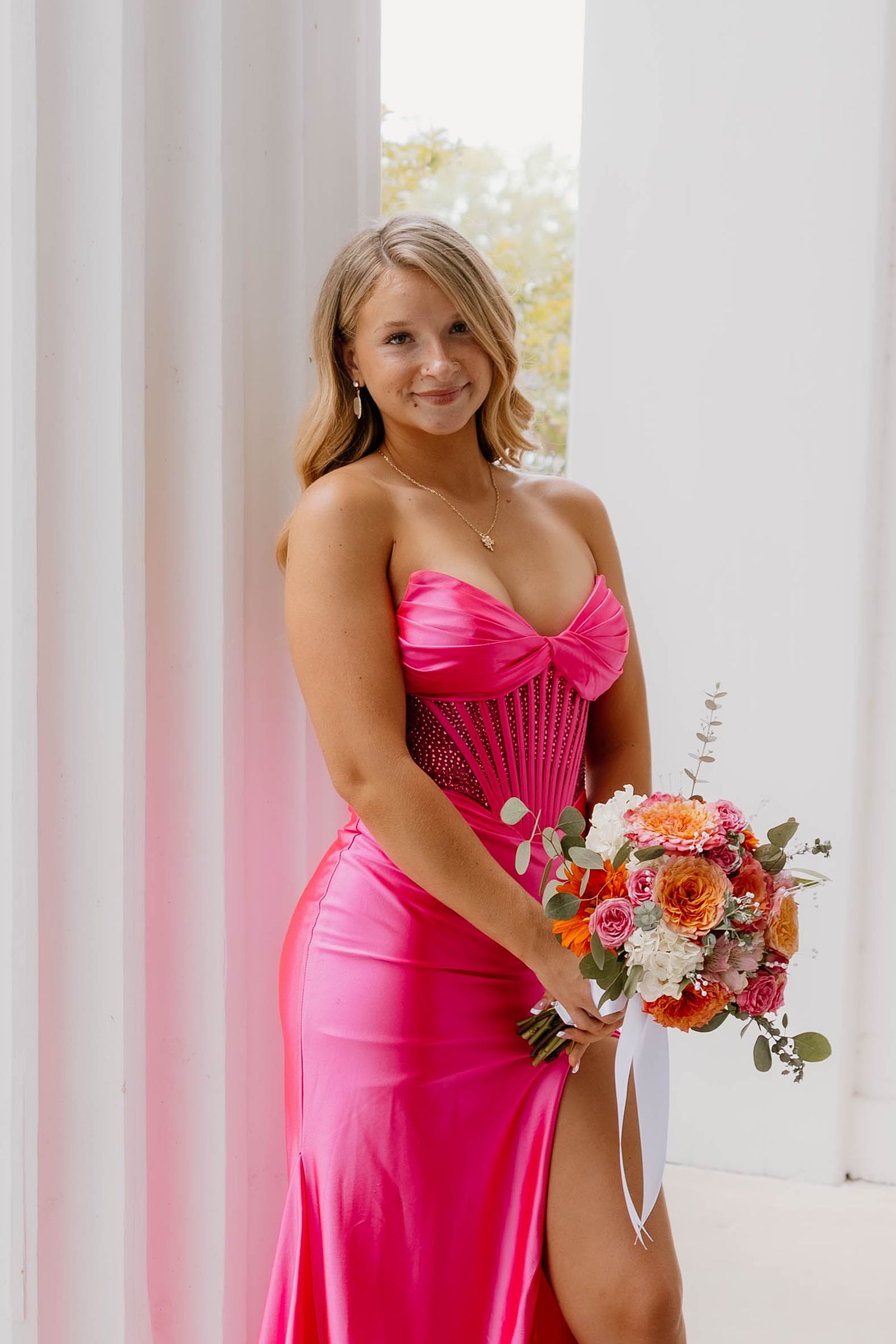 Woman in pink strapless satin gown holding colorful floral bouquet, posing between white columns.