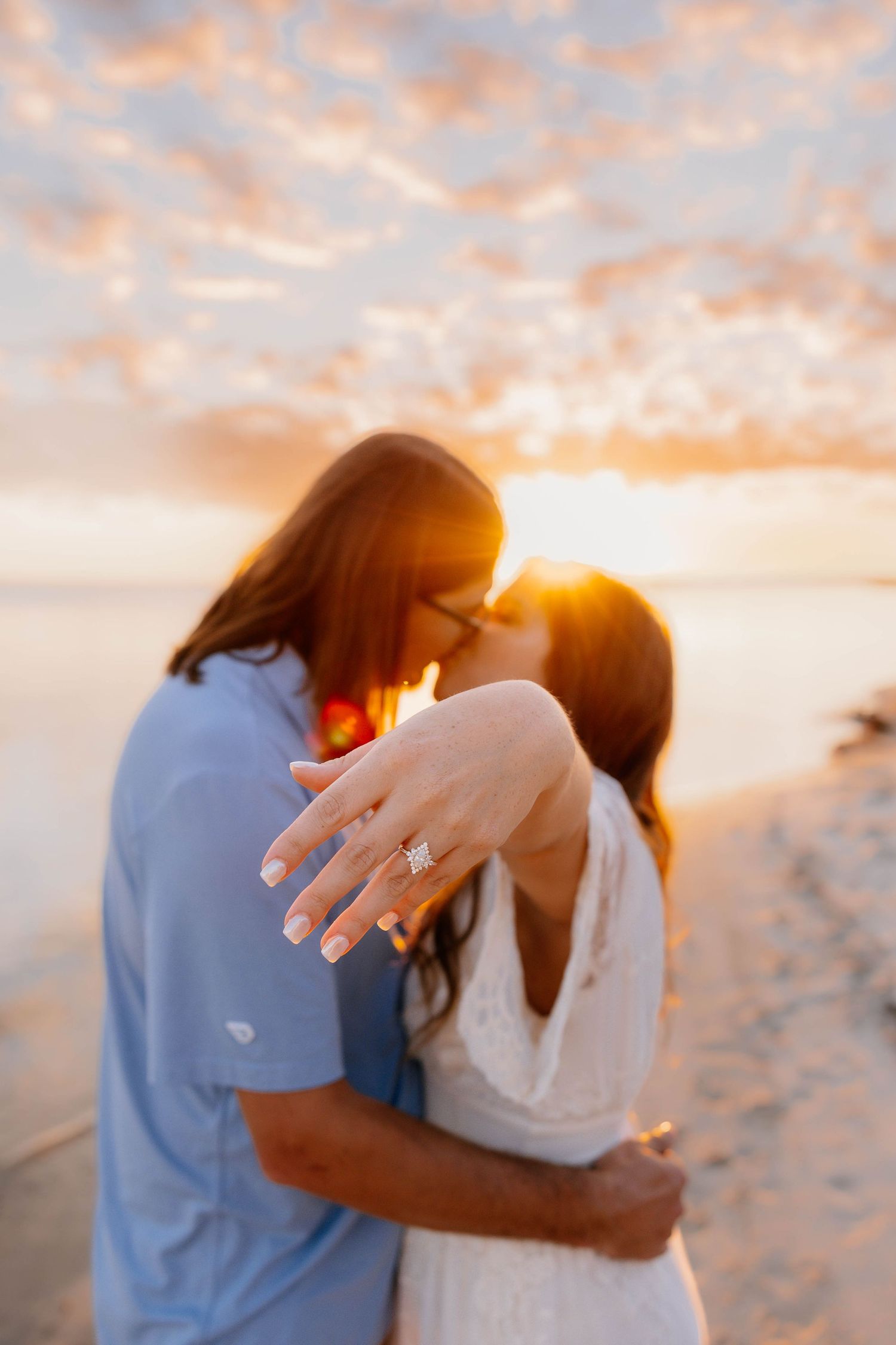 A couple embraces on a beach at sunset while showing off an engagement ring in golden light.