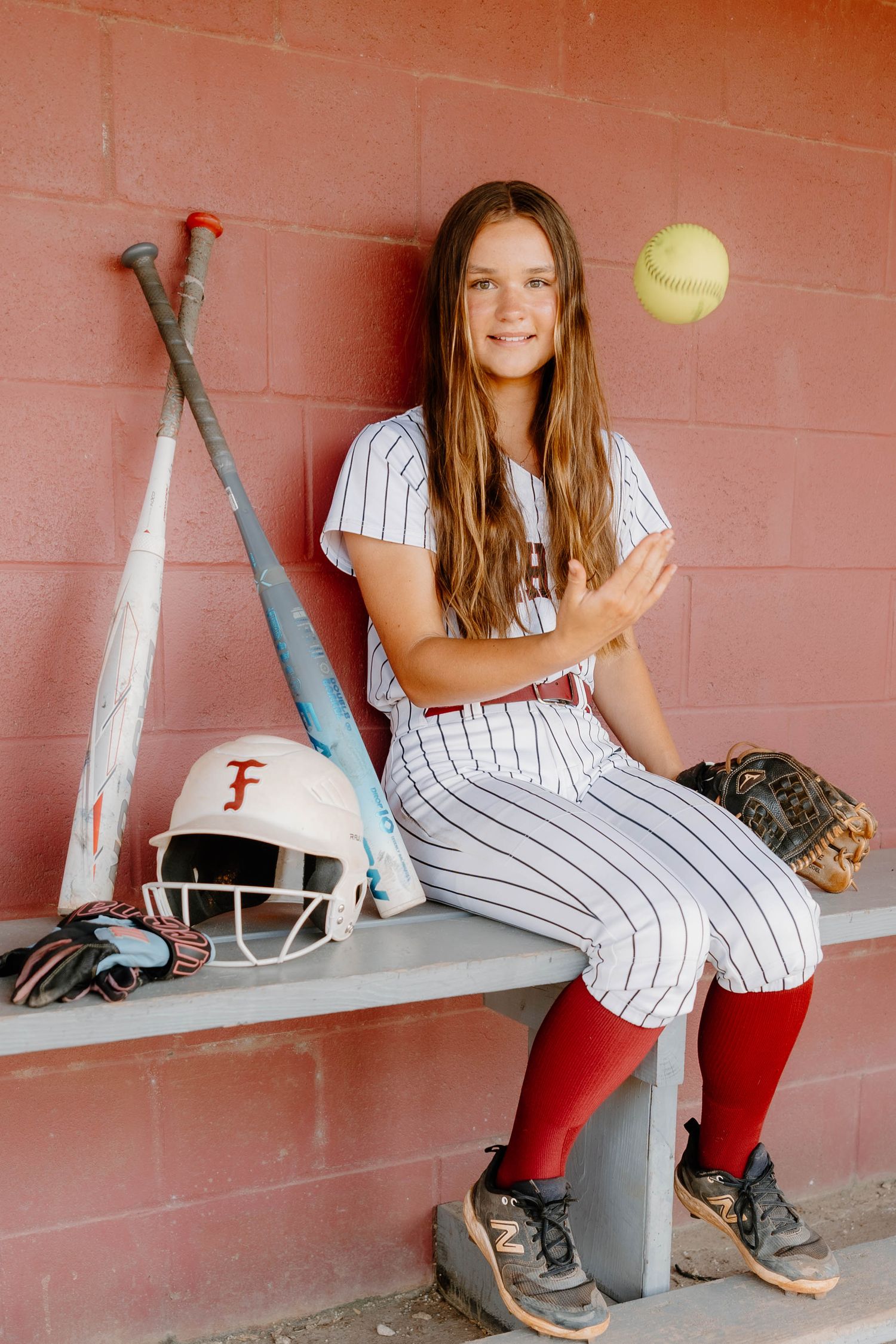 Young softball player in uniform sits in dugout, tossing a ball, with bat, helmet, and glove nearby.