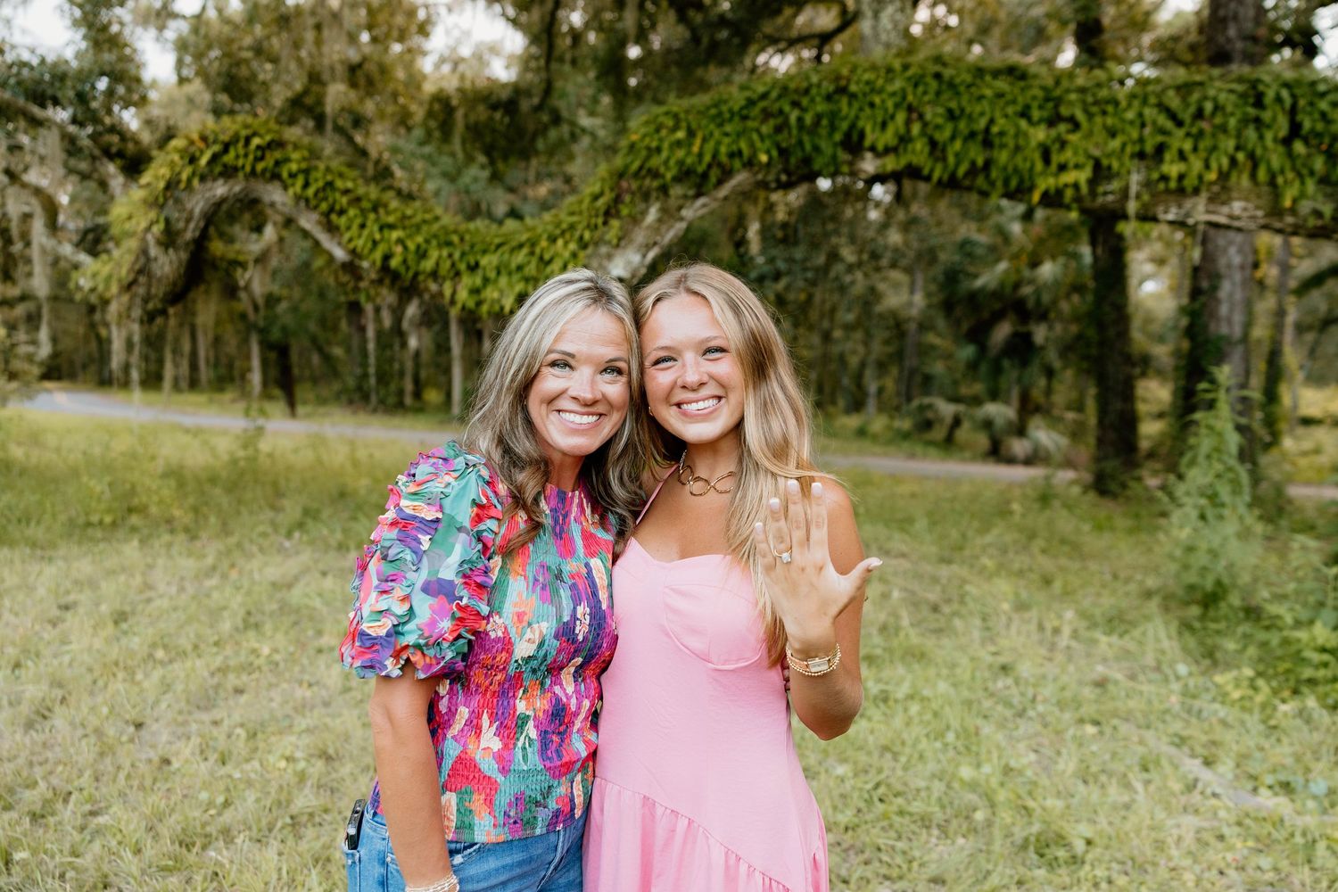 Two smiling women in pink outfits pose outdoors in a lush green park with moss-covered trees.