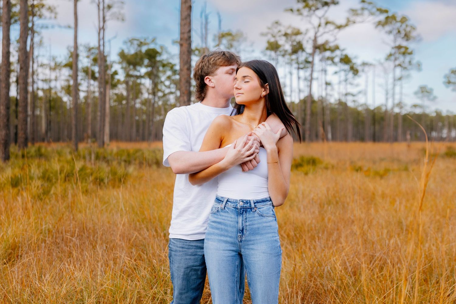 A couple embraces romantically in a golden field surrounded by tall pine trees under a cloudy sky.