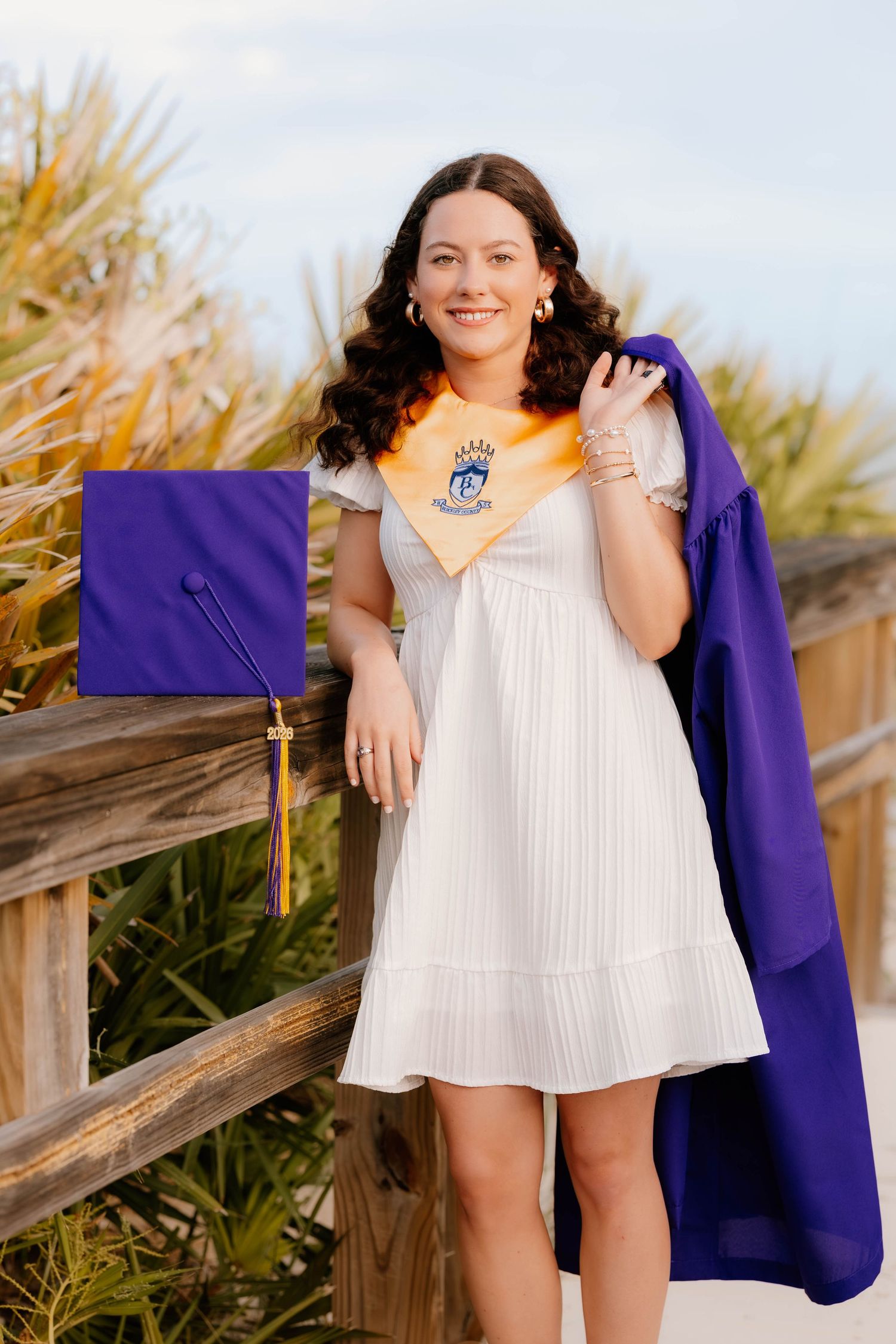 Smiling graduate in white dress holds purple robe and gold stole outdoors near wooden fence and tropical plants.
