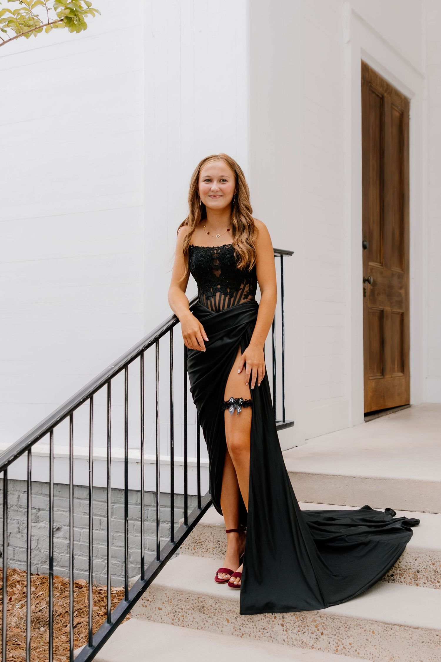 Young woman in elegant black formal gown with lace bodice and high slit poses on white staircase for prom photos.